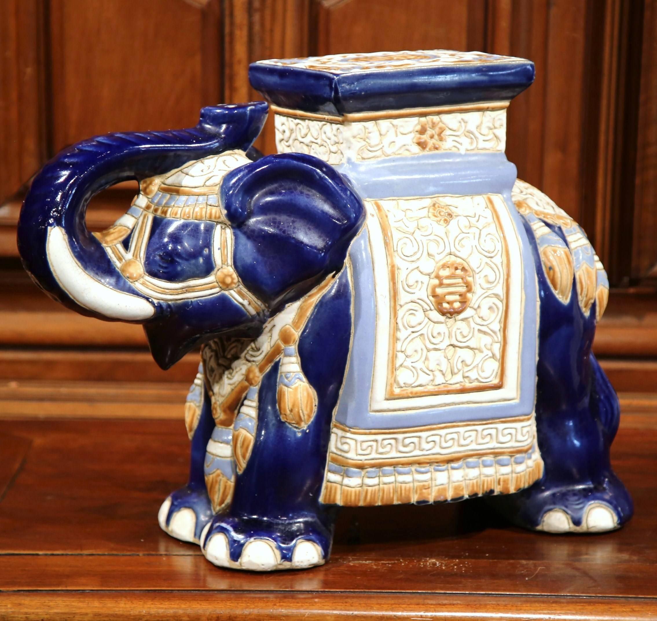 This interesting, porcelain garden seat was sculpted in France, circa 1950. The vintage piece is in the shape of an elephant raising his trunk, which is heavily decorated in oriental finery. The mammal has a square pedestal on top, and is