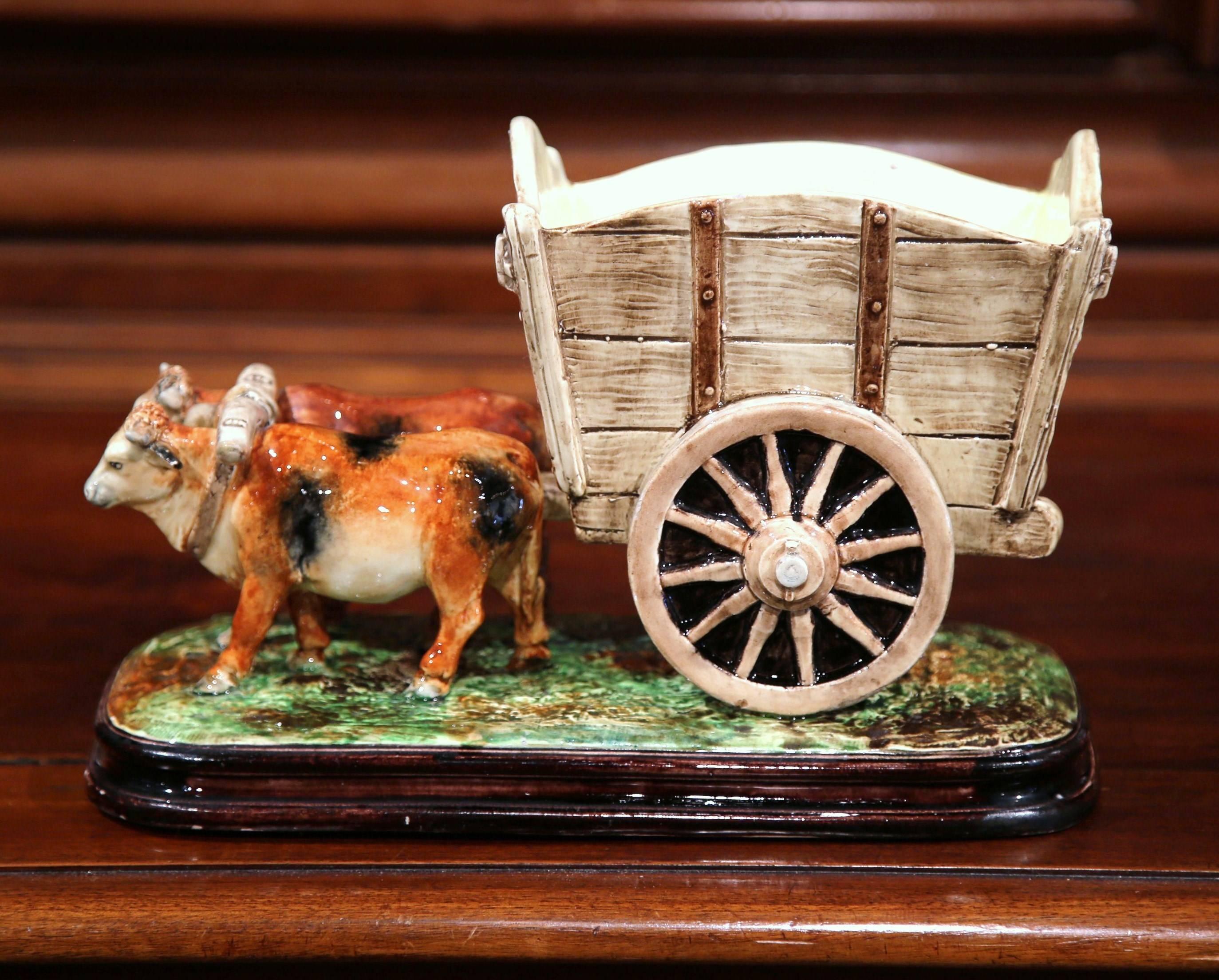 This colorful, antique Majolica sculpture composition depicts a Classic farm scene with cows pulling a cart. Created in France circa 1870, the ceramic jardinière features a pair of bovines joined together by a yoke, pulling a large wagon on wheel.