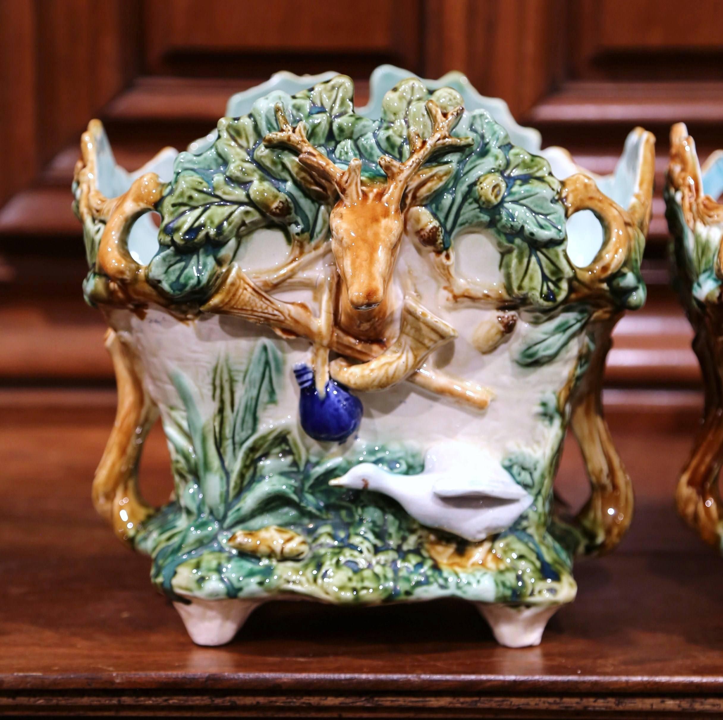 This pair of colorful, hand-painted Majolica jardinières were sculpted in France, circa 1880. The antique planters are almost square in shape. Each porcelain cache pot features exquisitely rendered hunting motifs on all four sides, which include