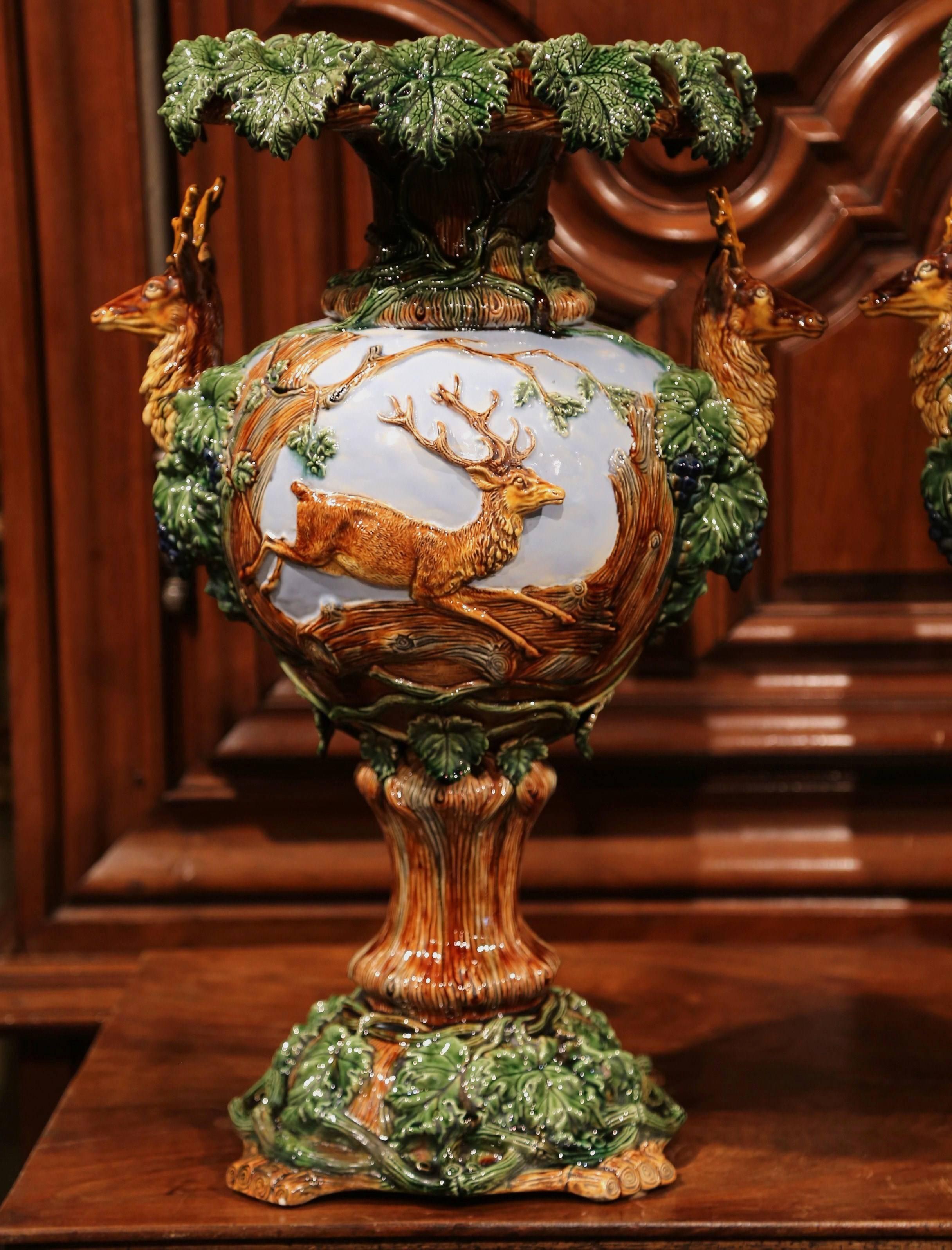This monumental pair of antique majolica vases was crafted in France, circa 1890. Each of the colorful vases features low relief stags running on the front with sculptural, high relief deer head trophies with antlers on the sides. The vases are