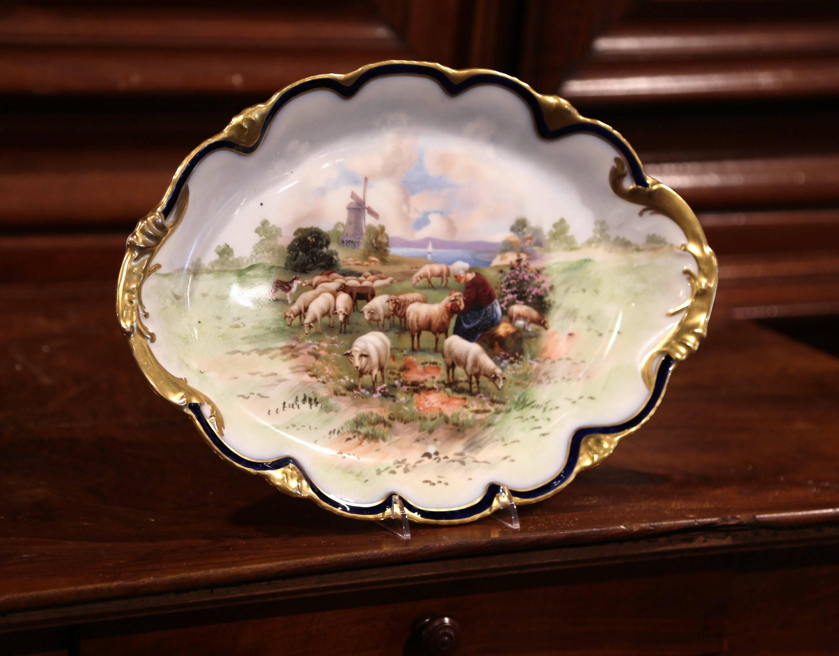 Beautiful antique platter from France crafted, circa 1920 the colorful plate features a hand-painted pastoral scene with a shepherdess, sheep, dog and wind mill in the background. The oval wall hanging plate with gold leaf around the trim is stamped