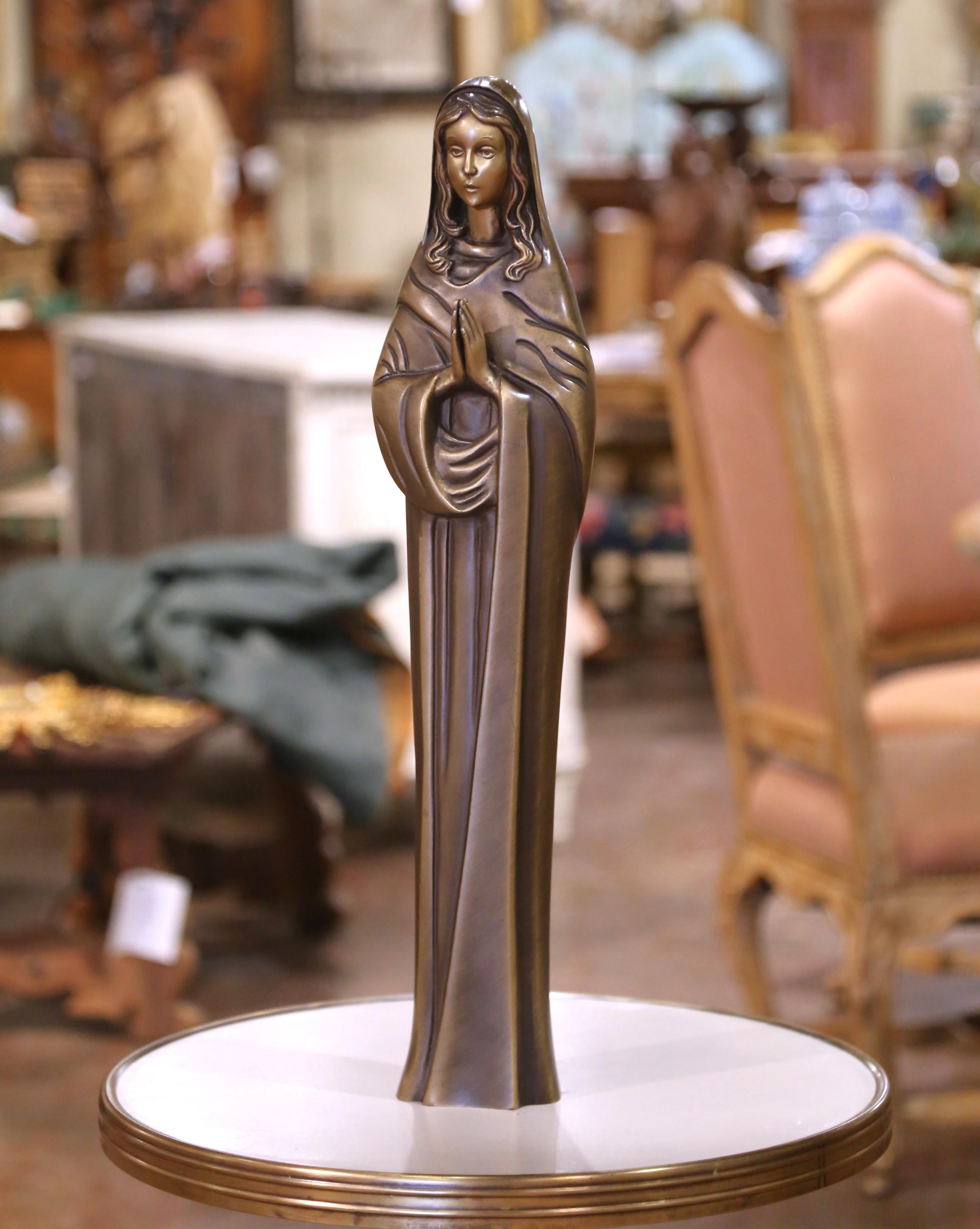 Moulée en France vers 1960, cette élégante sculpture en bronze du milieu du siècle représente la Vierge Marie dans une dévotion paisible, les mains pressées dans la prière et les yeux doucement baissés. La forme stylisée et allongée reflète