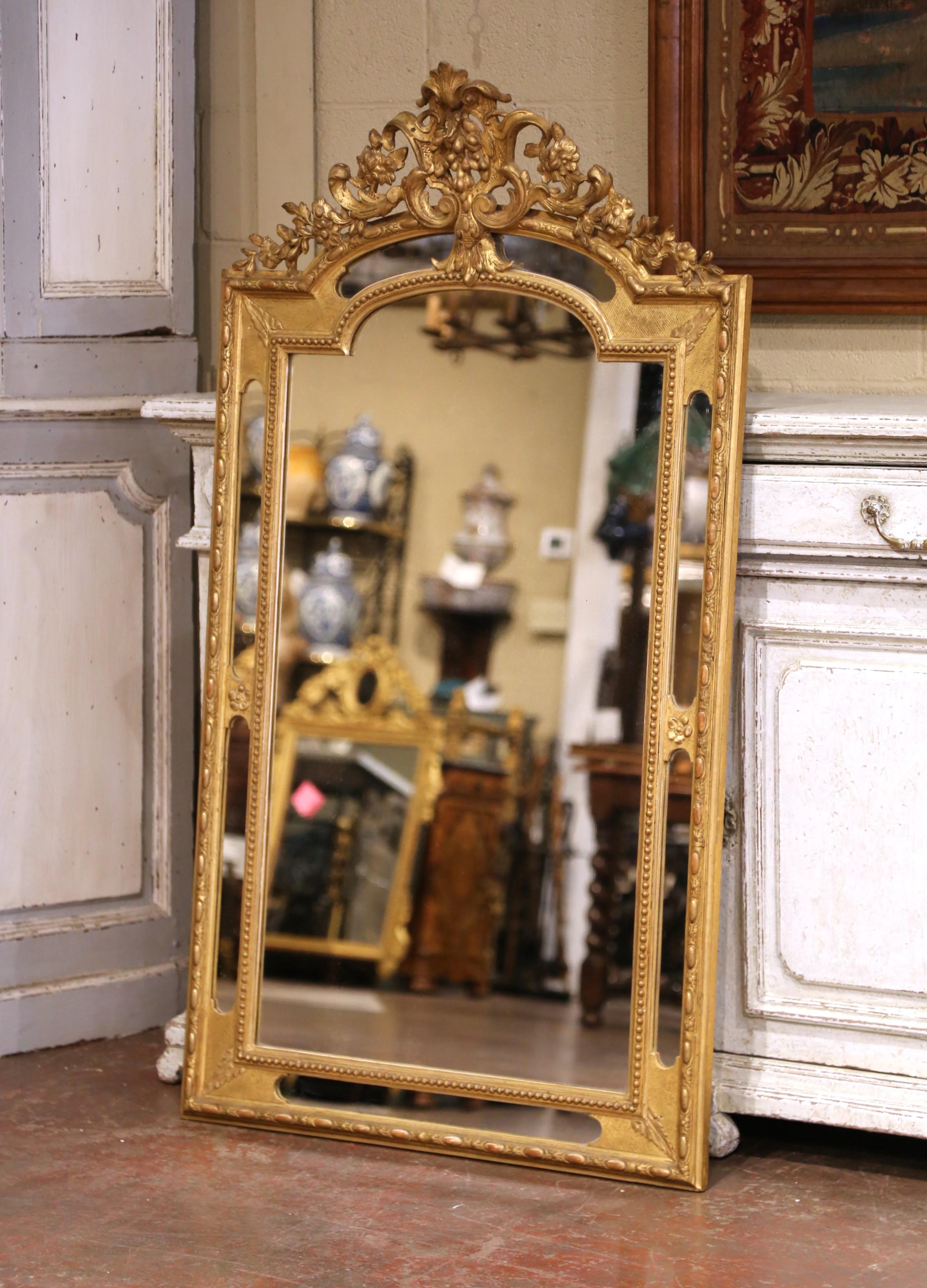 De casi metro y medio de altura, este antiguo espejo de pared se fabricó en Francia hacia 1860 y capta la refinada elegancia del estilo Luis XVI. El marco de madera dorada tallada está rematado con una elaborada cresta con una palmeta central,