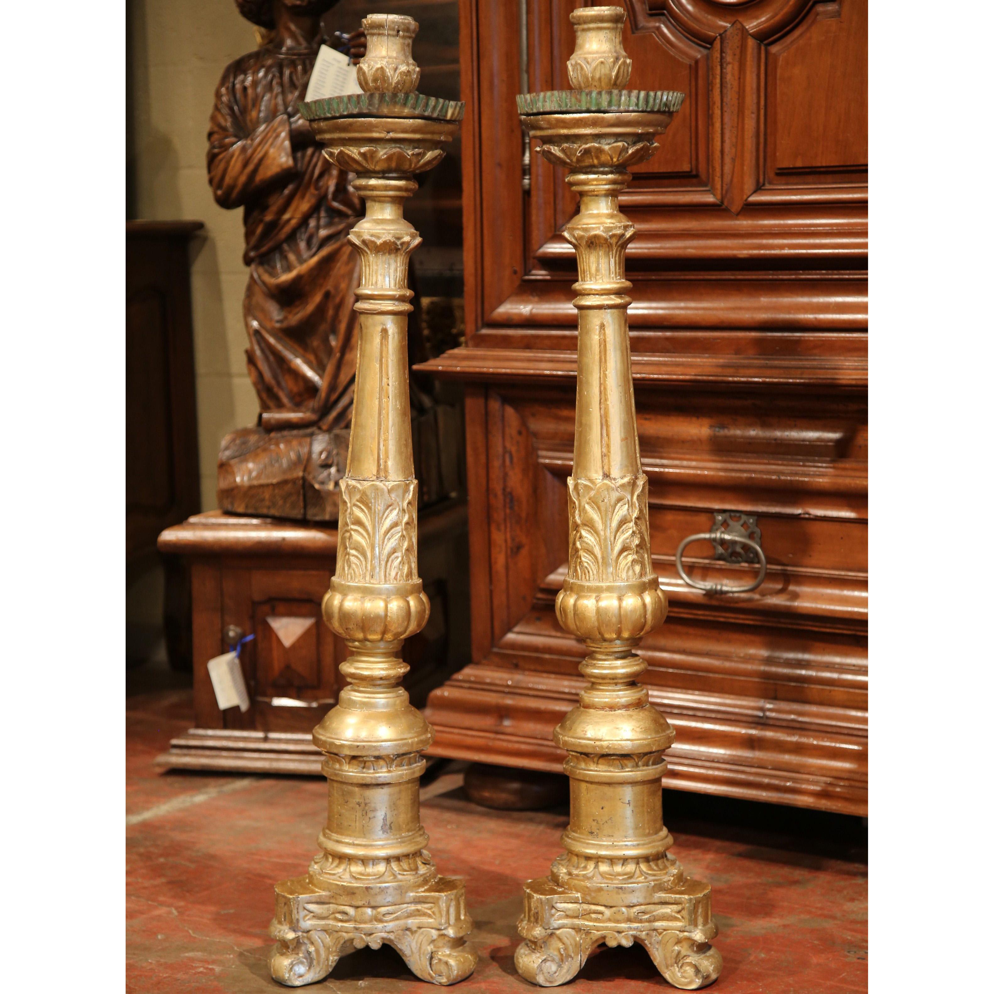 This tall and decorative pair of antique candlesticks was hand-carved in France circa 1780 in the Louis XV style. Each piece features a richly ornamented front with fluted columns, stylized acanthus leaves, and gadrooned detailing, resting on a