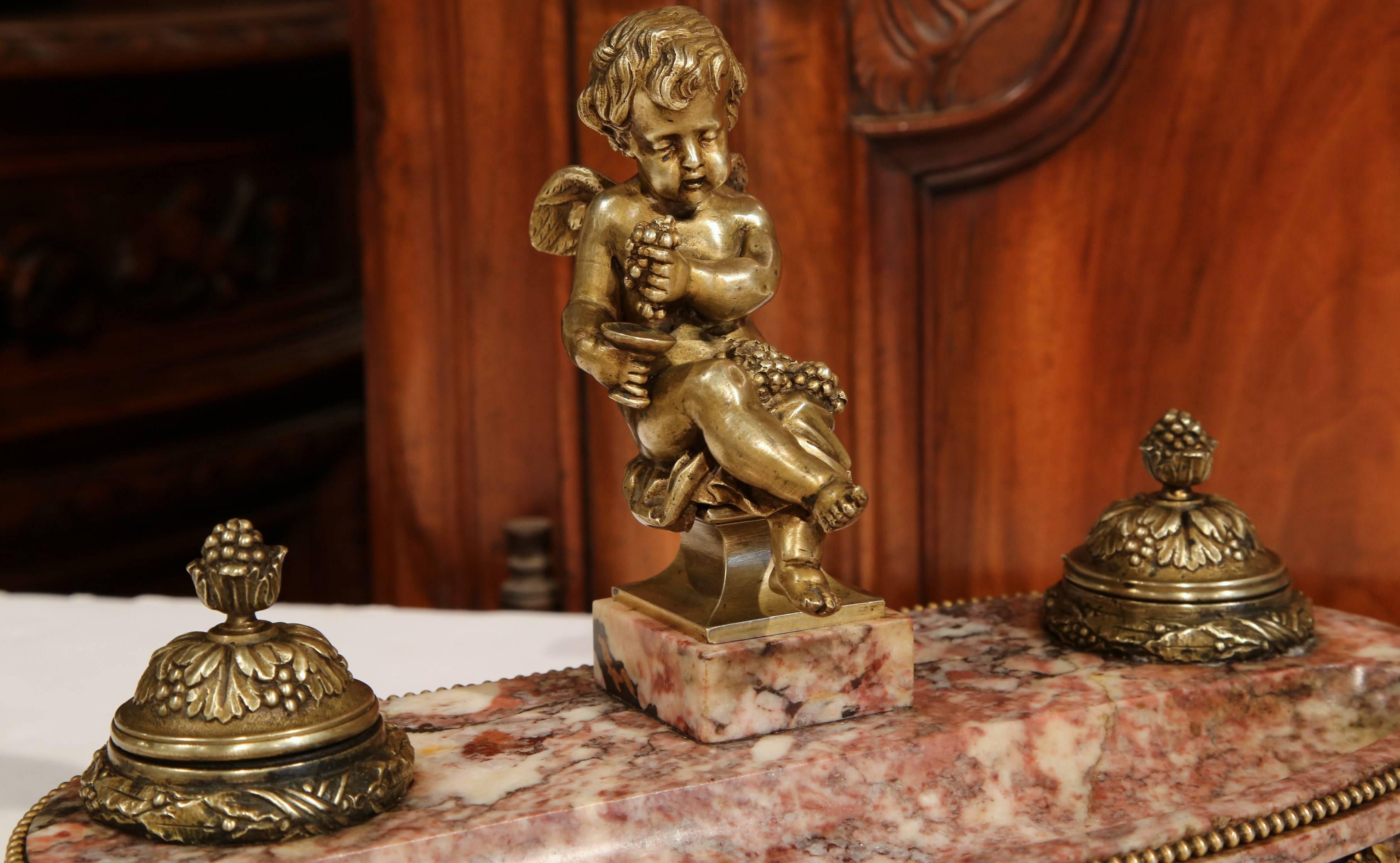 This fine, antique bronze and marble inkwell was crafted in France circa 1860. The inkwell is decorated with a seated putti with wings holding grapes. On either side of the cherub, both ink holders are lined with their original glass containers (one