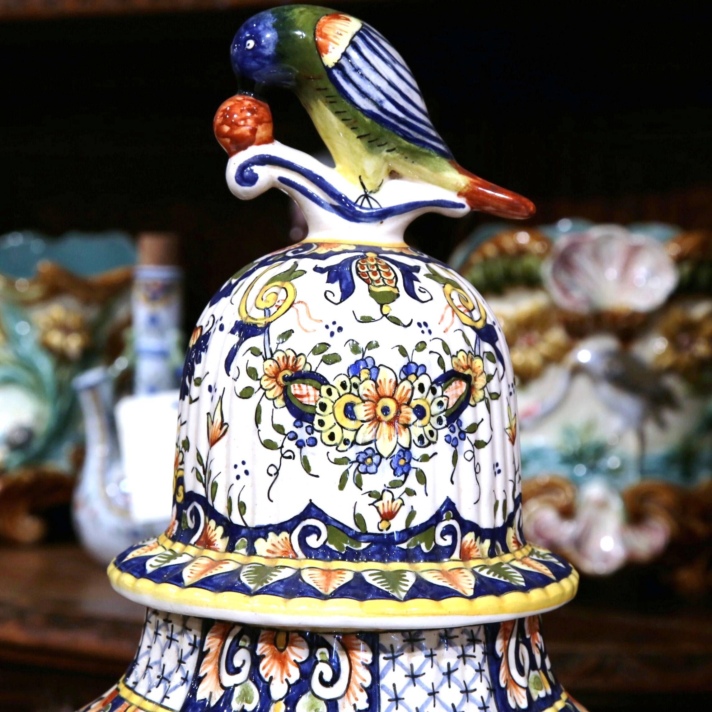 This large, colorful, hand-painted potiche with lid was sculpted in Normandy, France. The tall antique vase features rich colors with floral motifs in a blue, red and yellow palette on a white background. The lid has a parrot pecking at a cherry,