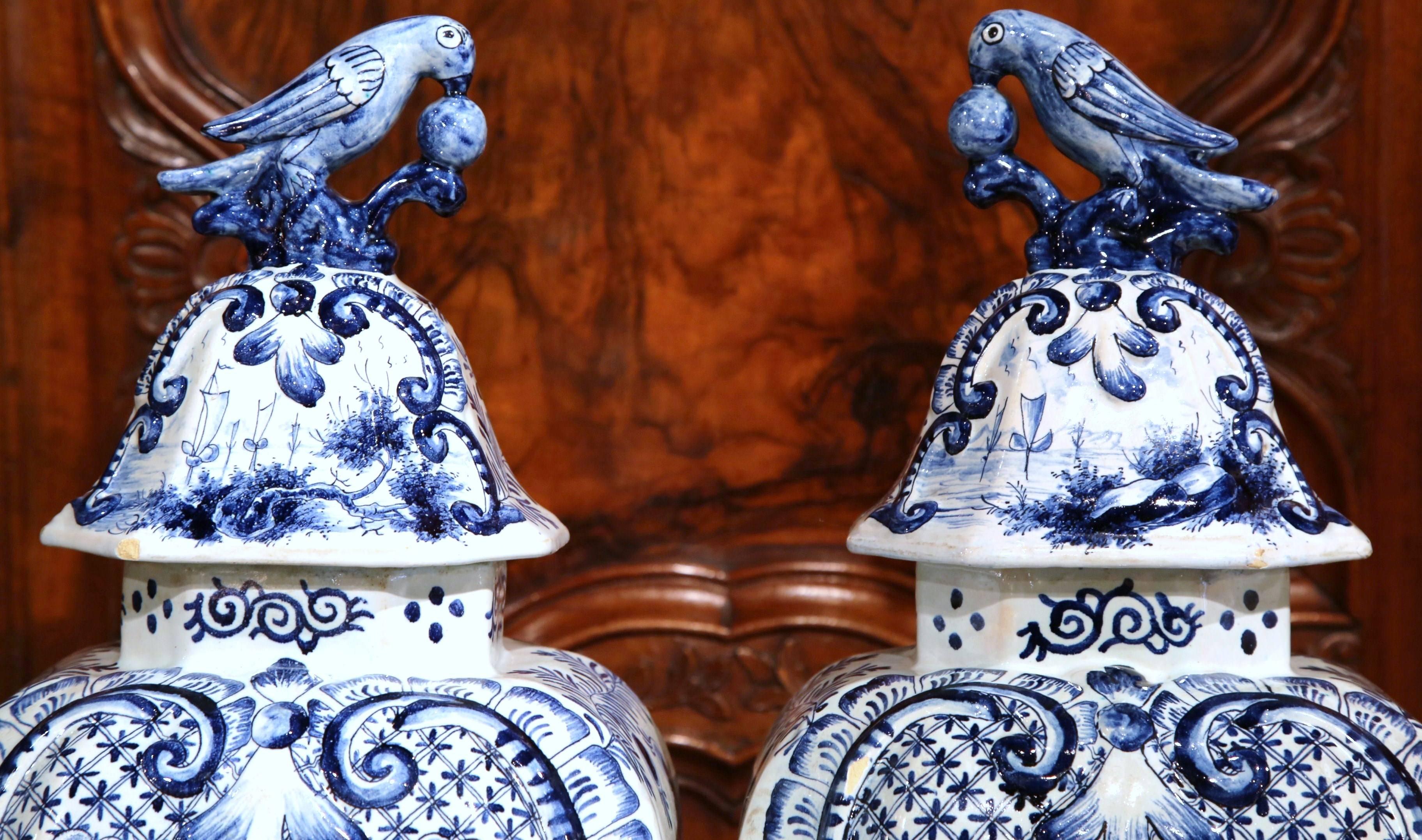 This Classic pair of blue and white delft vases from 1880 depict finely painted, romantic scenes. The base and shoulders of the vases are decorated with an intricate diamond lattice design. On top of the vase, the lids are embellished with a