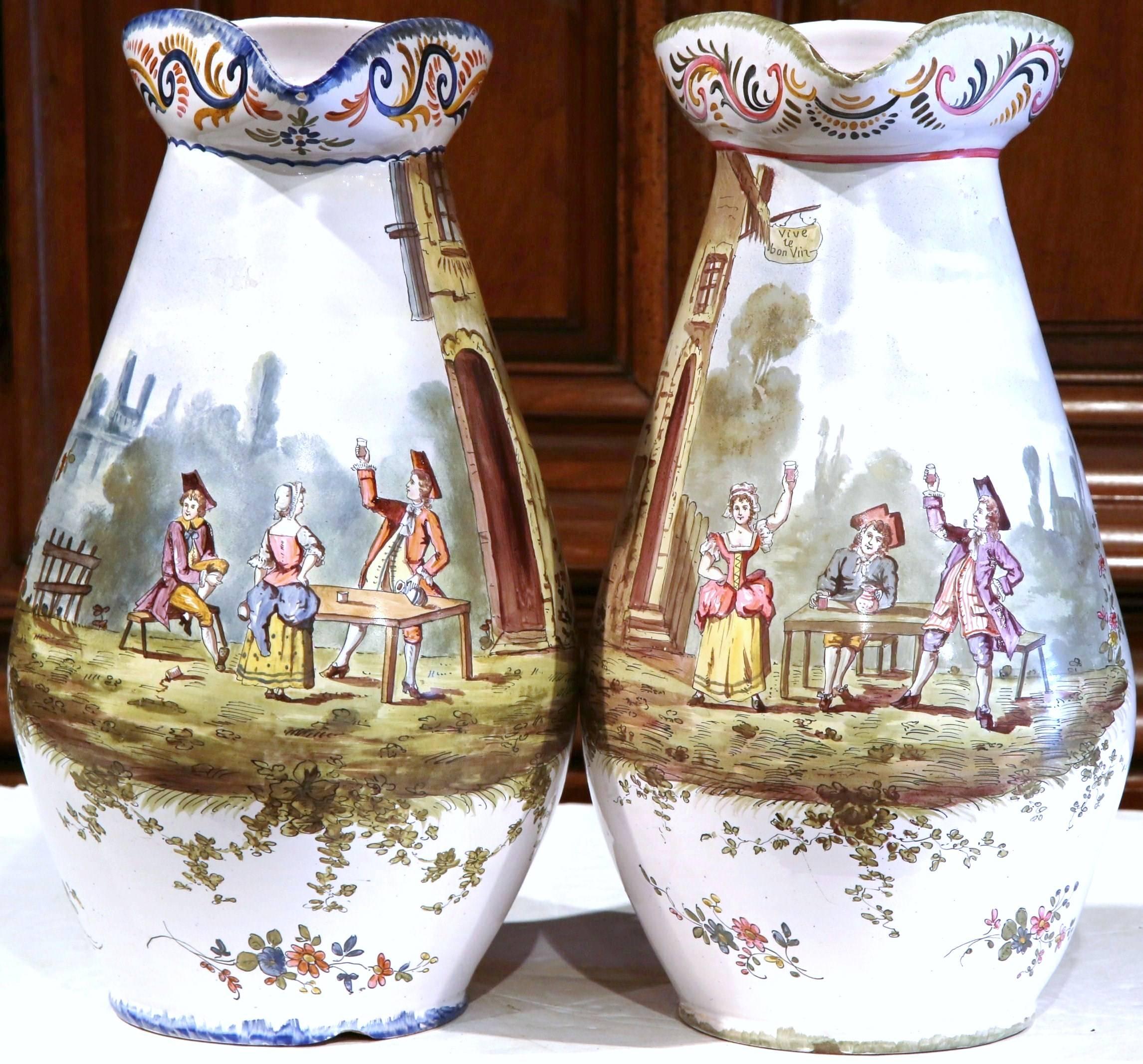 This tall pair of antique, porcelain pitchers was created in Burgundy, France circa 1880. The rustic amphoras feature hand-painted tavern and pastoral scenes with people sitting around a table while drinking wine and toasting to each other. One
