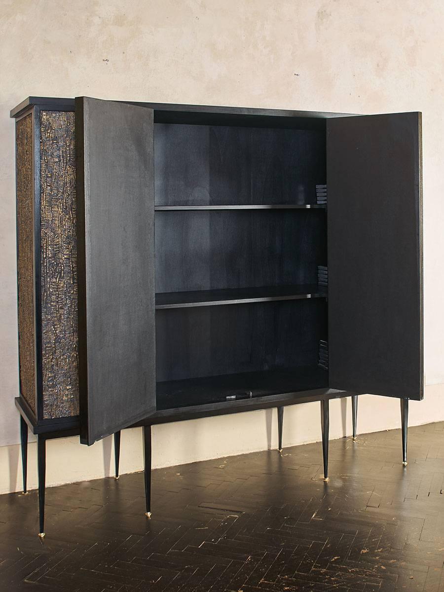 Black cabinet decorated with resin and brass powder panels, fixed shelves inside, black steel base with natural brass details.