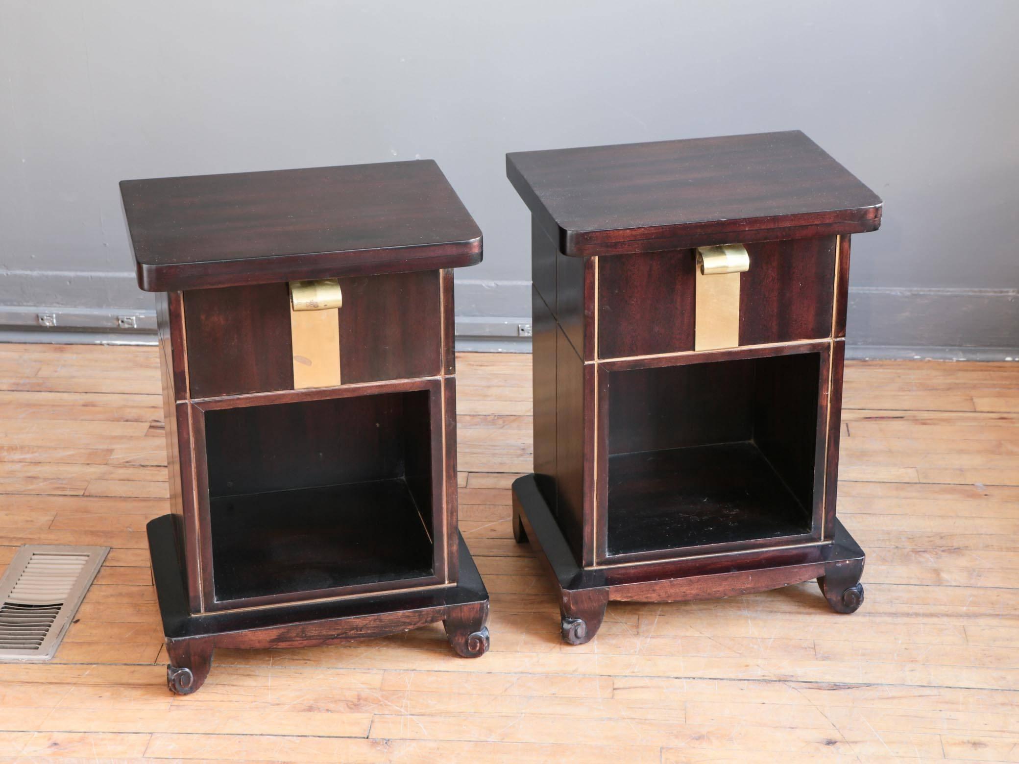 A rare pair of Art Deco nightstands or side tables in mahogany by John Stuart of New York, circa 1930s or 1940s. Each featuring one deep drawer with a sculptural, solid brass pull above an open shelf. Incised gilt decoration and scroll feet add