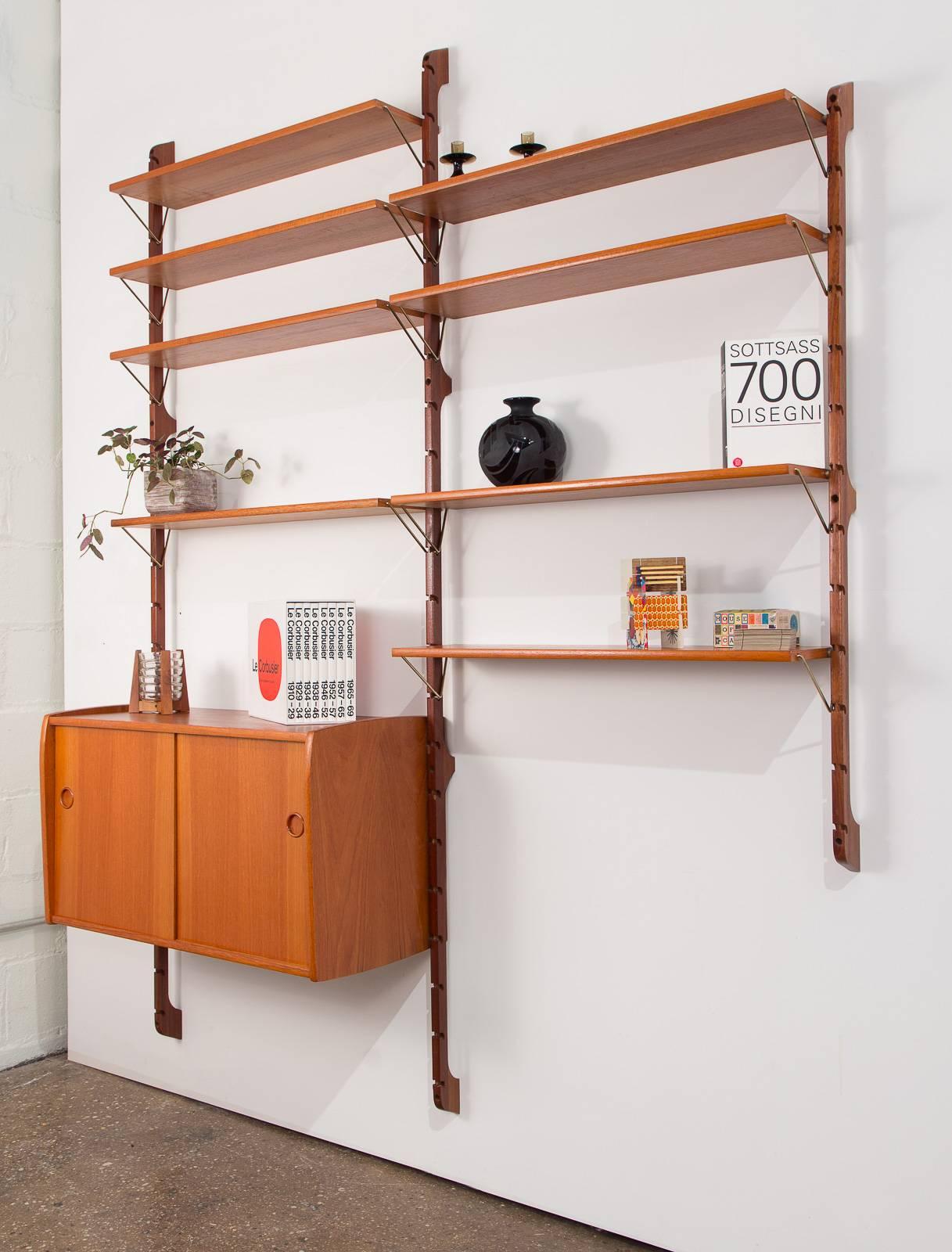 Beautiful 1960s Norwegian wall unit for Blindheim Møbelfabrik. This modular three-bay teak unit comes with eight shelves and one sliding-door cabinet. The shelves can be installed with the angular brass brackets above or below the shelves. Cabinet