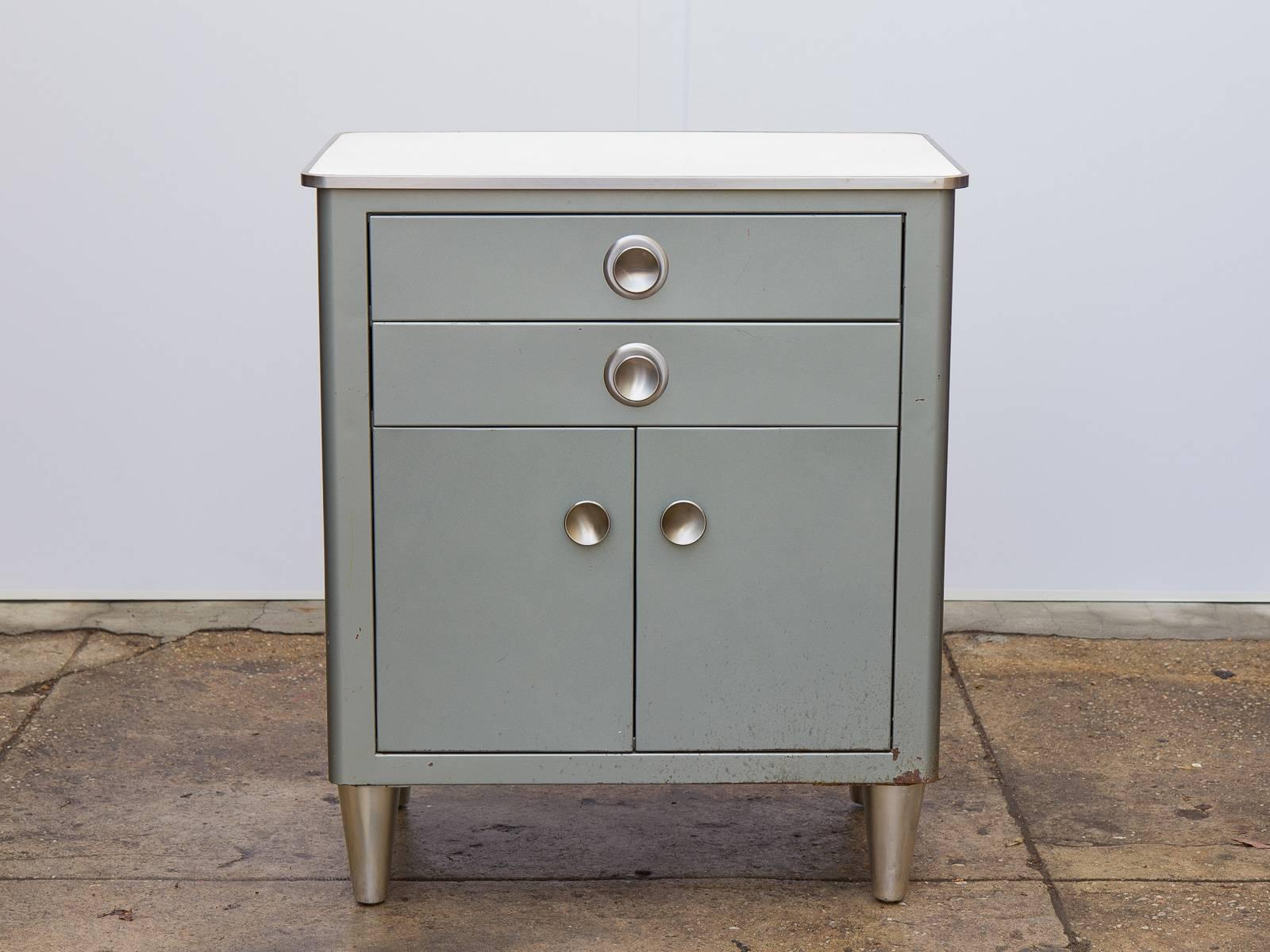 Small Modern Industrial display cabinet manufactured by American Metal Furniture, Inc. This hefty—yet sleek—Norman Bel Geddes-inspired steel cabinet sports round disc pulls on the drawers and cabinet doors. Charming linoleum speckled ivory