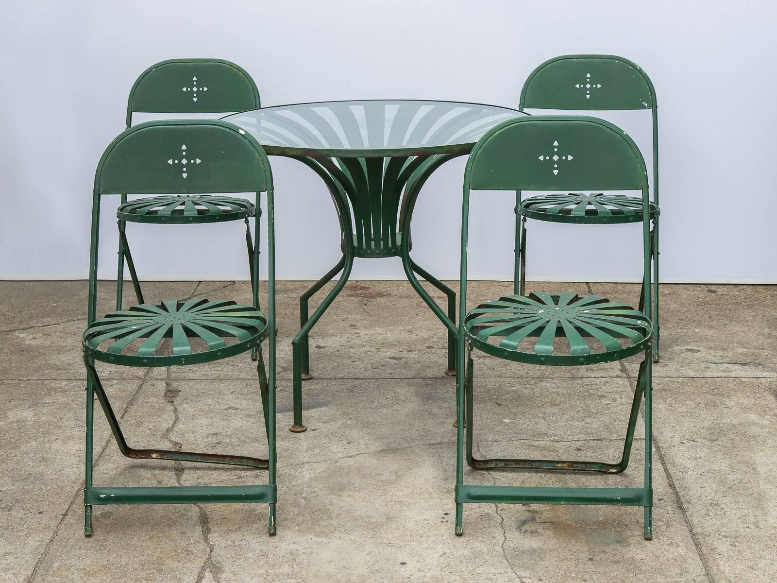 Four cut-out wrought iron folding chairs and glass table in green enamel. This set is in the style of Francois Carre's, 1930s Sunburst outdoor furniture featuring the same sunburst seats and table base form. All pieces are in good vintage condition