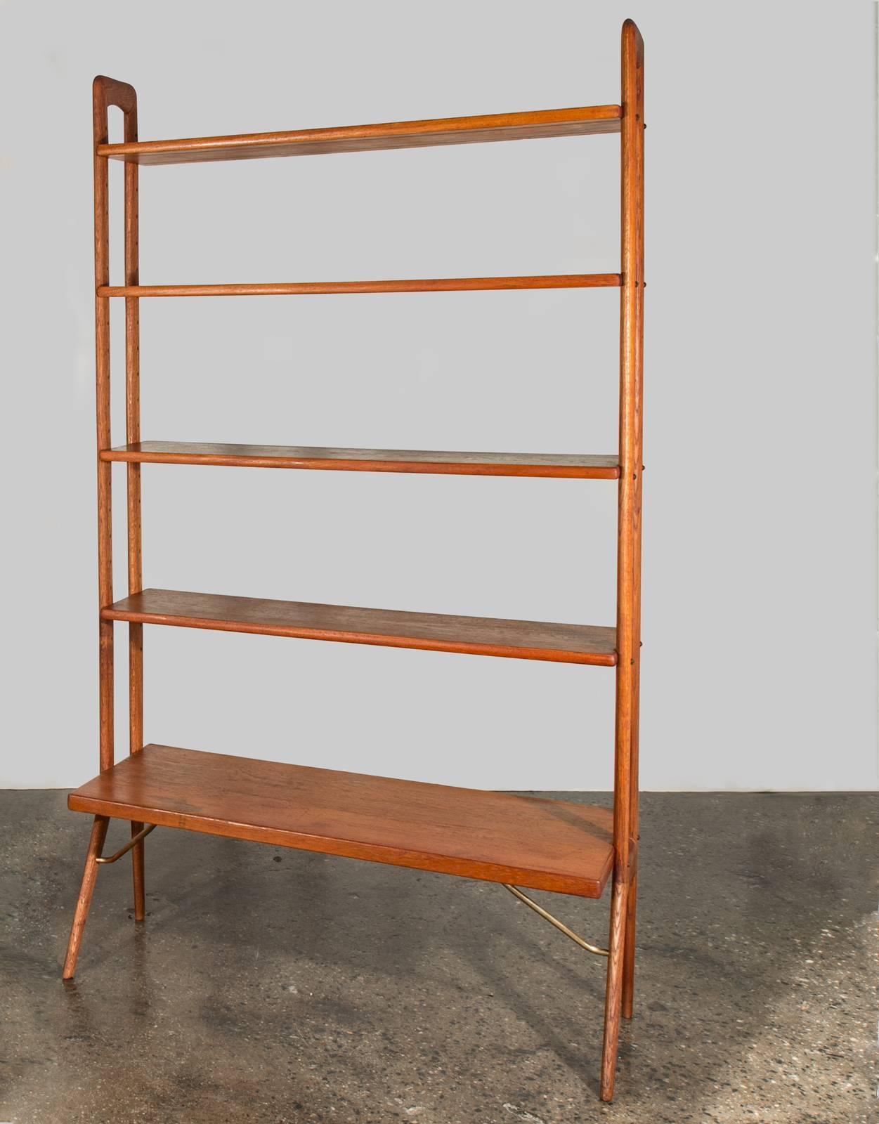 Graceful teak bookcase designed by Kurt Østervig. The shelves are adjustable, and the holes for repositioning form an integral part of the aesthetic appeal.

The wide shelf is deep enough to hold vinyl records and is supported by handsome brass