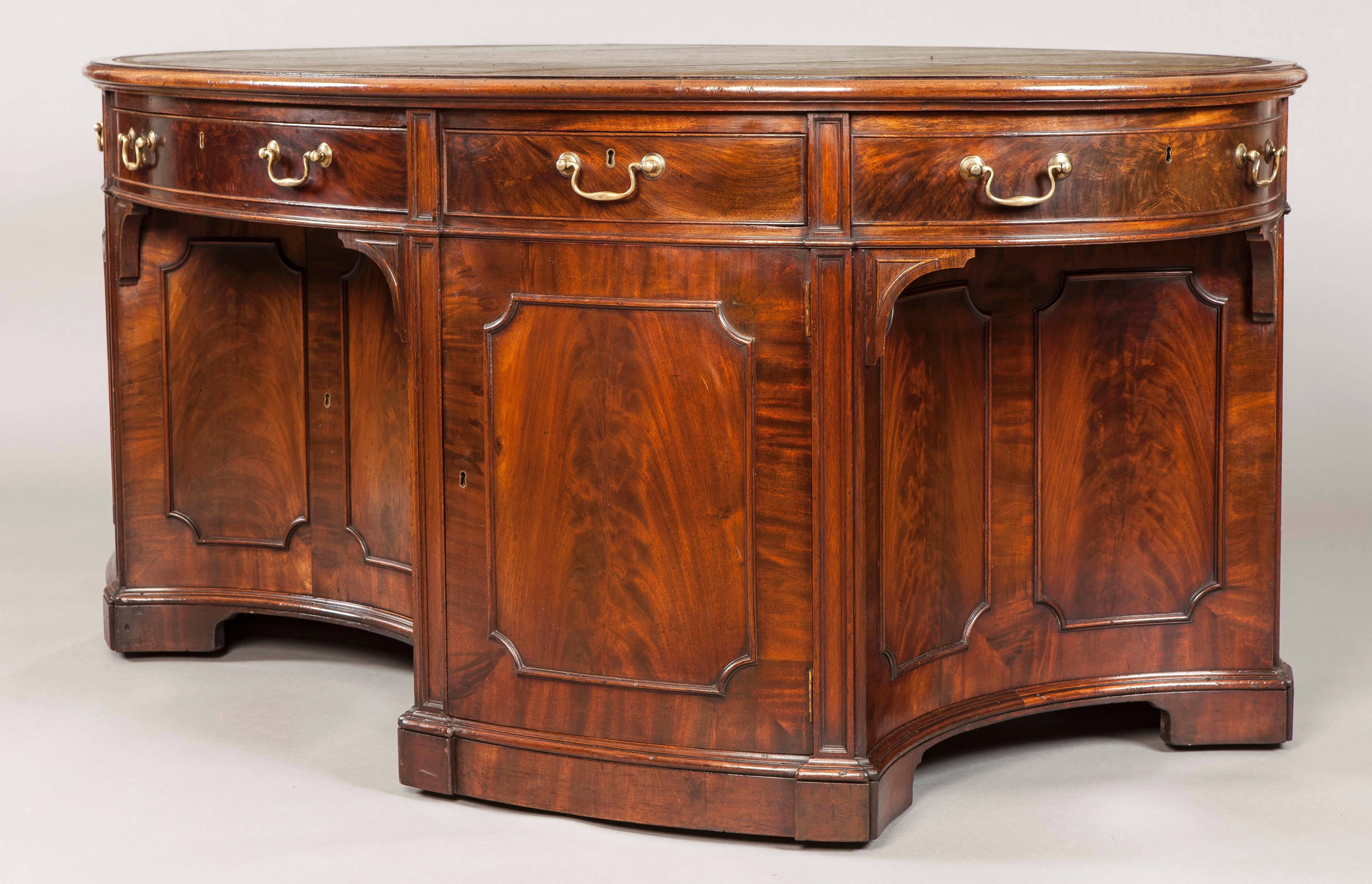 An elliptical library table after a design by Thomas Sheraton.

Constructed in a well figured mahogany; of double sided free standing form, having cupboards adorned with mouldings flanking the kneeholes, set within a series of vertical stiles and