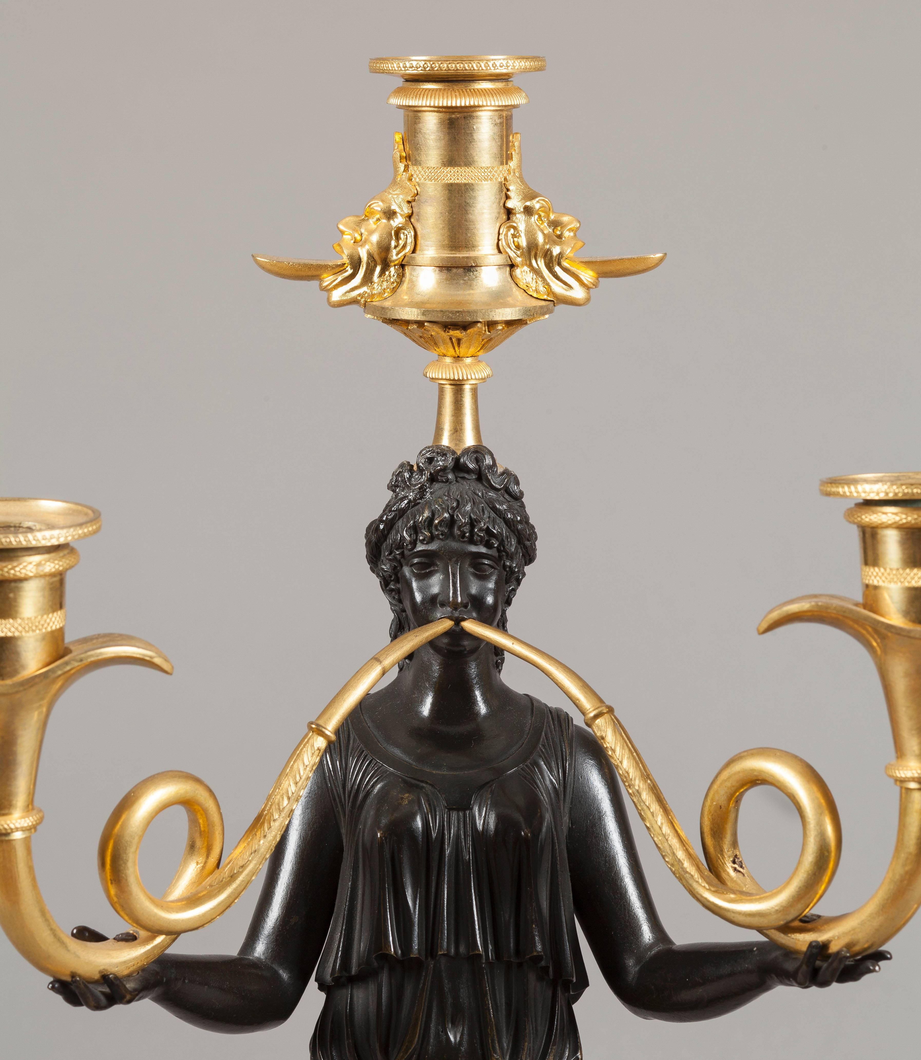 
Constructed in two color gilt and patinated bronze; the feminine Grecian figures draped with diaphanous robes blowing bi-form trumpets of swept form, terminating in candleholders and capped with single candlesticks, dressed with grotesque masks,
