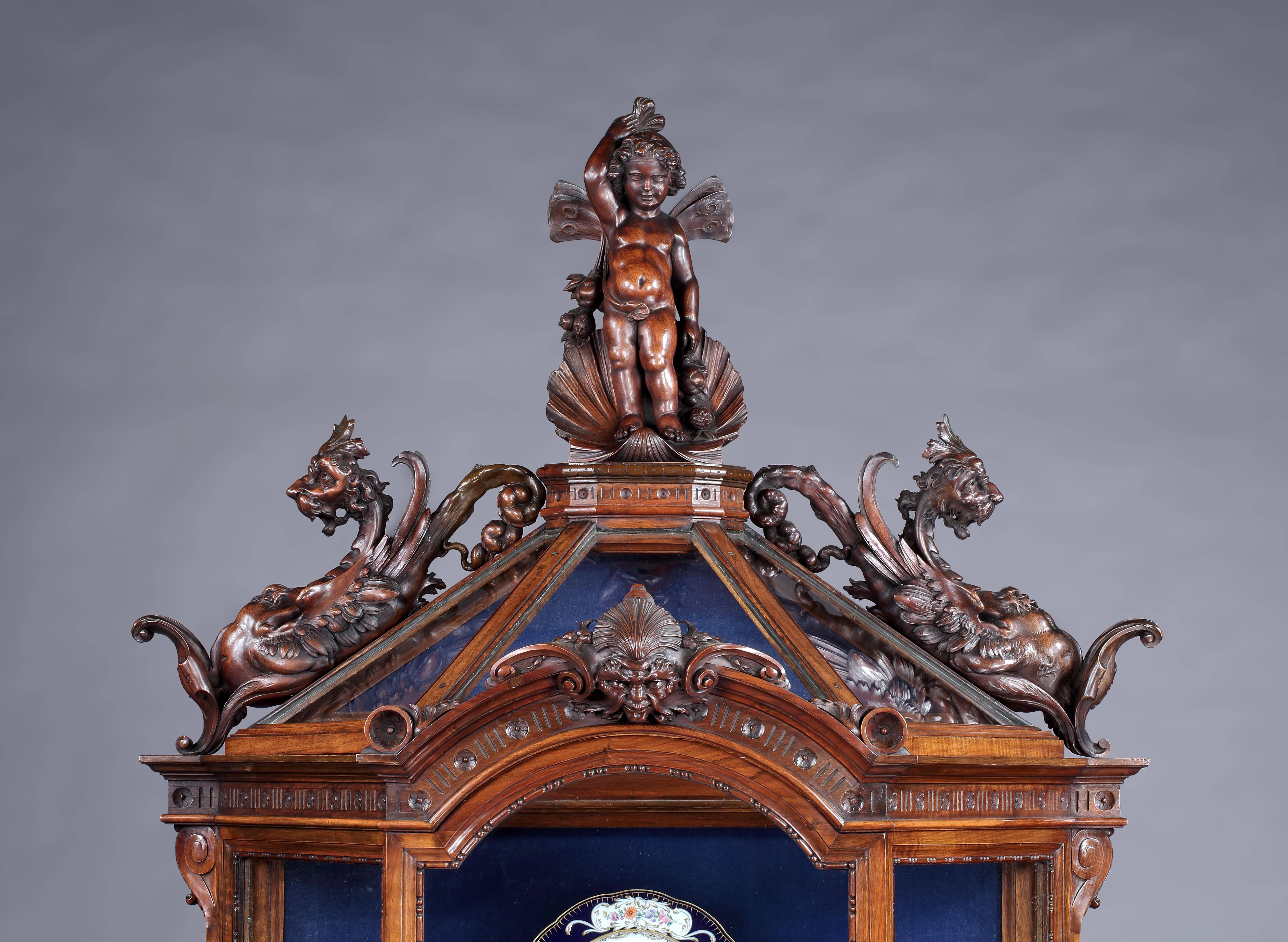An Italian carved walnut vitrine.

Constructed in carved walnut, rising from a serpentine ogee edged plinth base with a mirror back, having a central mythological winged figure flanked by turned columns supporting the glazed vitrine canted upper