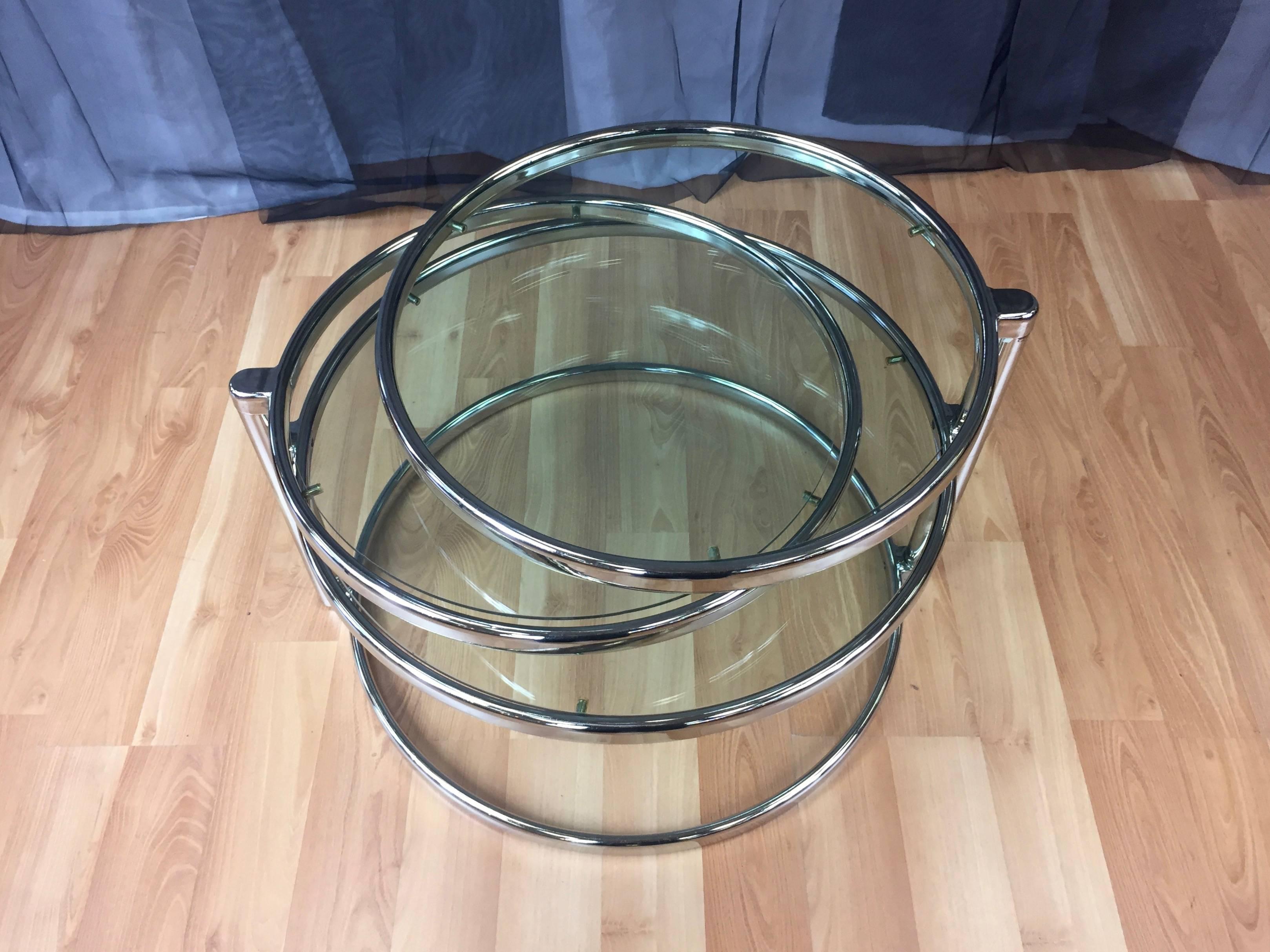 A Milo Baughman-style polished nickel and glass swiveling three-tiered round table.

Comprised of four stacked rings which include an open base, a glass-topped fixed tier and two glass-topped swiveling tiers. Transforms from cocktail table to