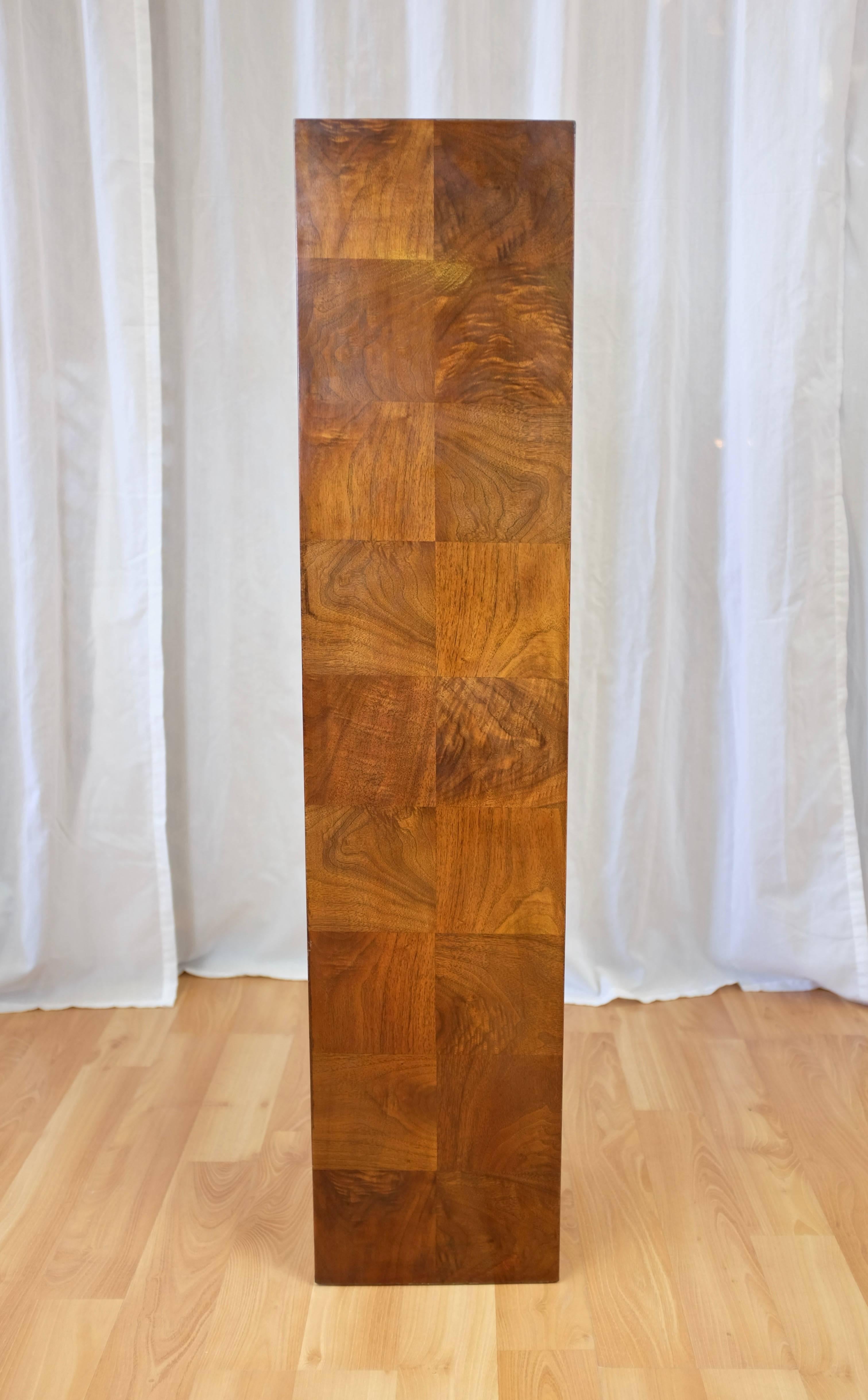 A circa-1970s tall walnut pedestal in the style of Milo Baughman.

Finished on all sides in lively figured crown cut walnut veneer squares with a bright, warm tone. An understated yet stately stage upon which to display a prized piece of art or