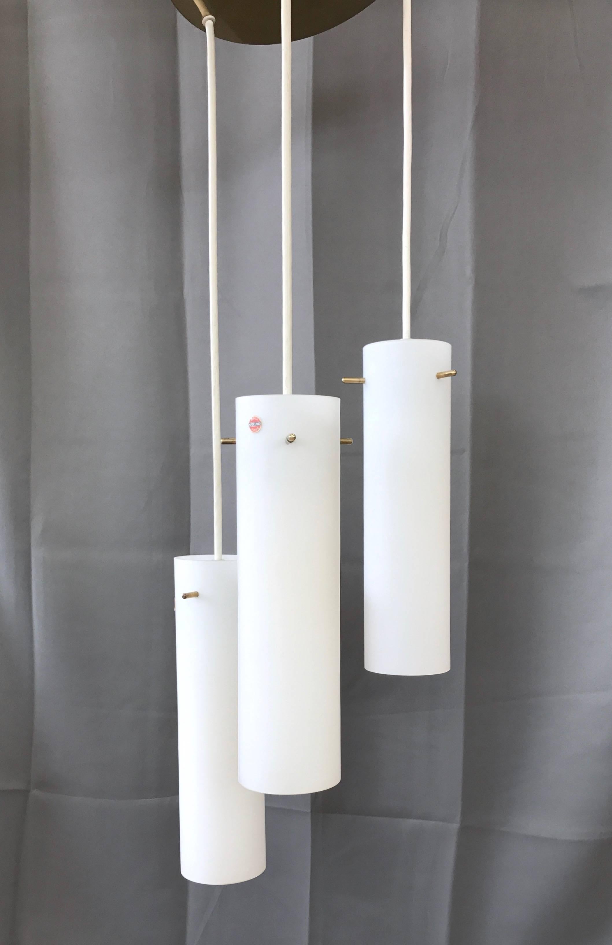 A Mid-Century triple pendant glass and brass ceiling light fixture by Hadeland Glasswverk of Norway.

A timeless example of Minimalist Scandinavian design, with three 14 in. H x 3.75 in. Dia frosted glass cylindrical shades hung in a staggered