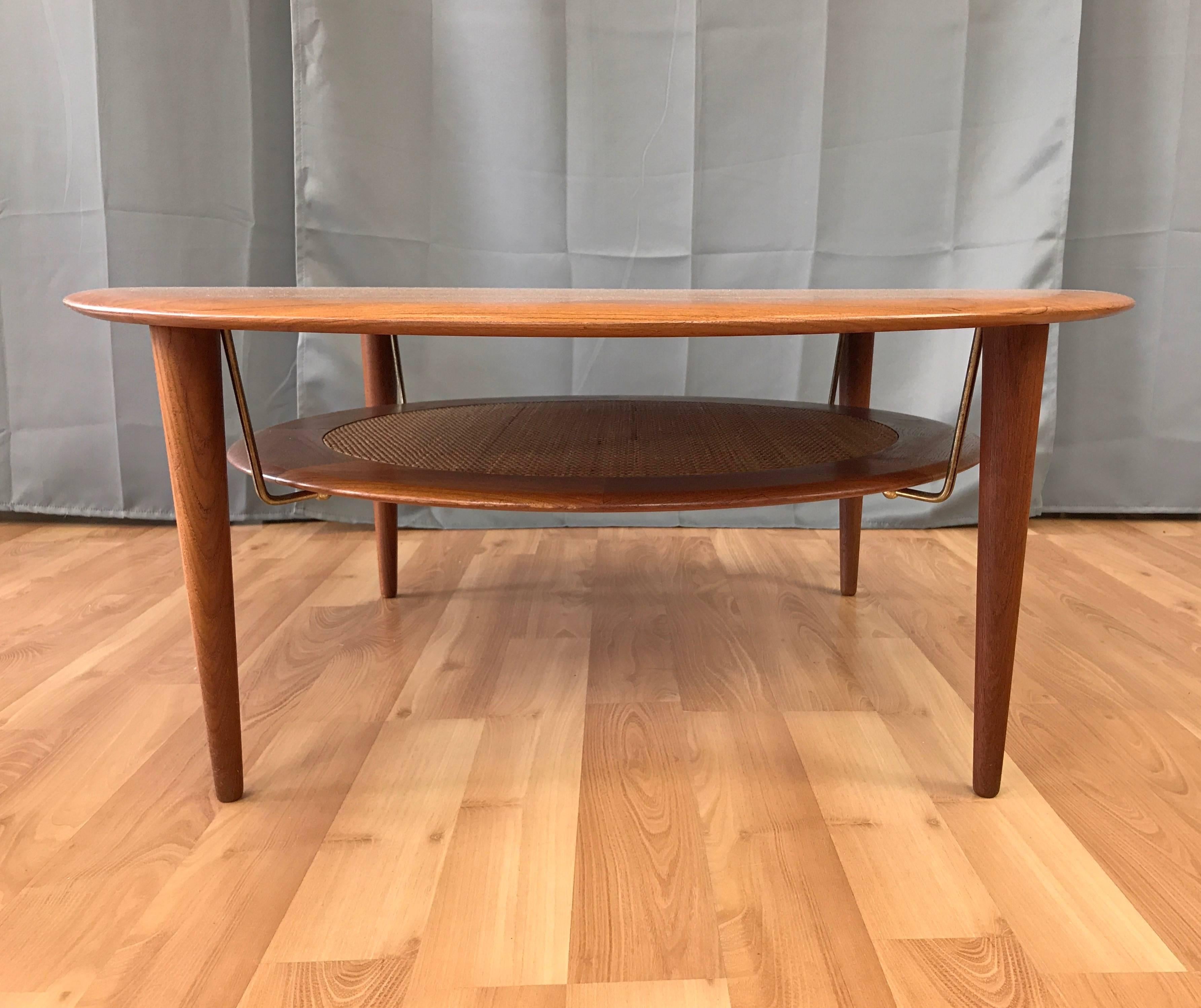 Handsomely distilled Danish modern design, with an expansive disc top and tapered legs handcrafted in solid teak. Floating lower tier features a sleek teak ring with original tightly woven rattan inset, and is suspended by four discreet brass rods.