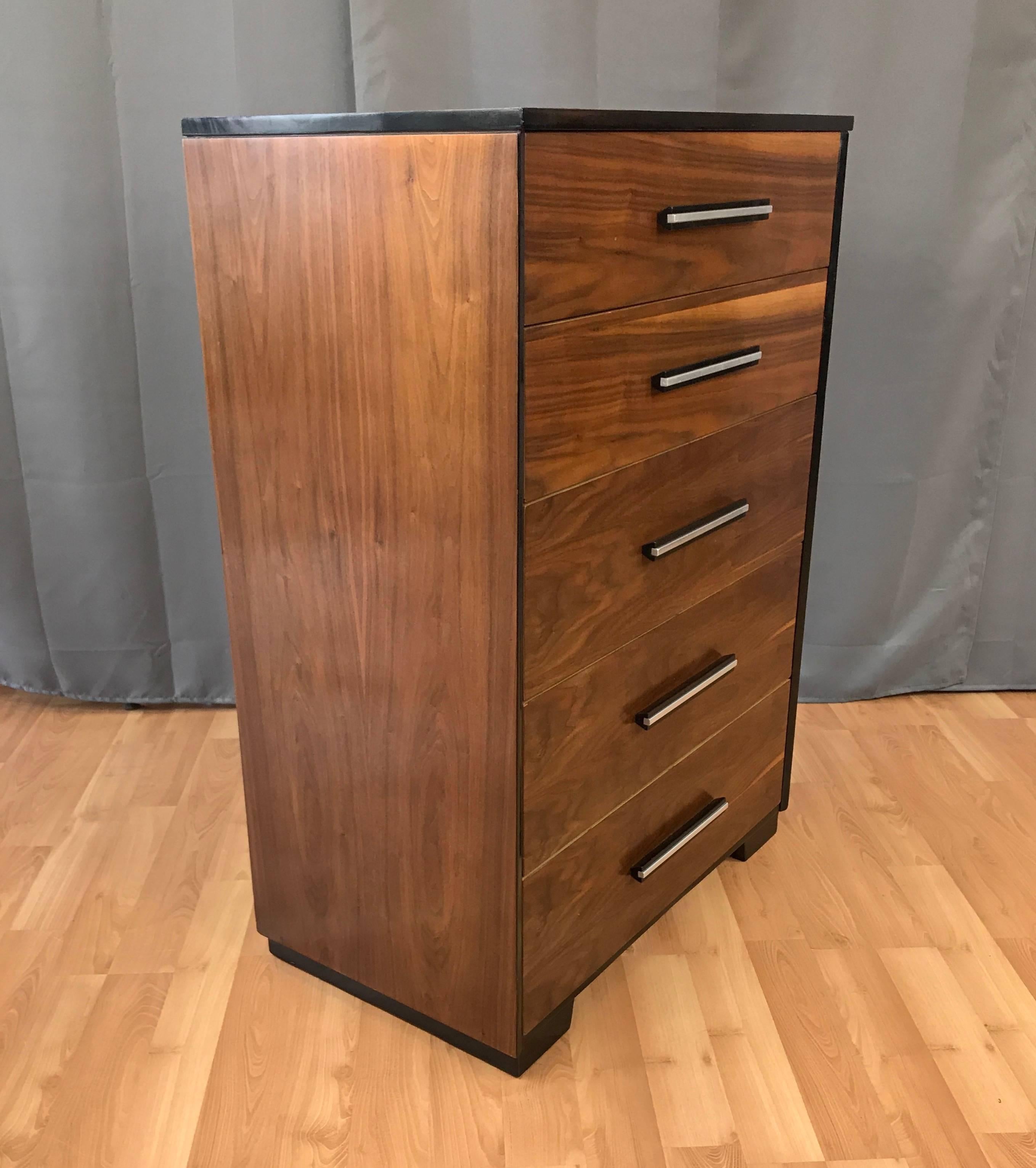 A midcentury walnut five-drawer highboy dresser designed by Raymond Loewy for Mengel Furniture.

Finished in very handsomely figured bookmatched walnut, with semi-gloss black lacquered trim that accents its clean, minimalist lines. Drawers feature