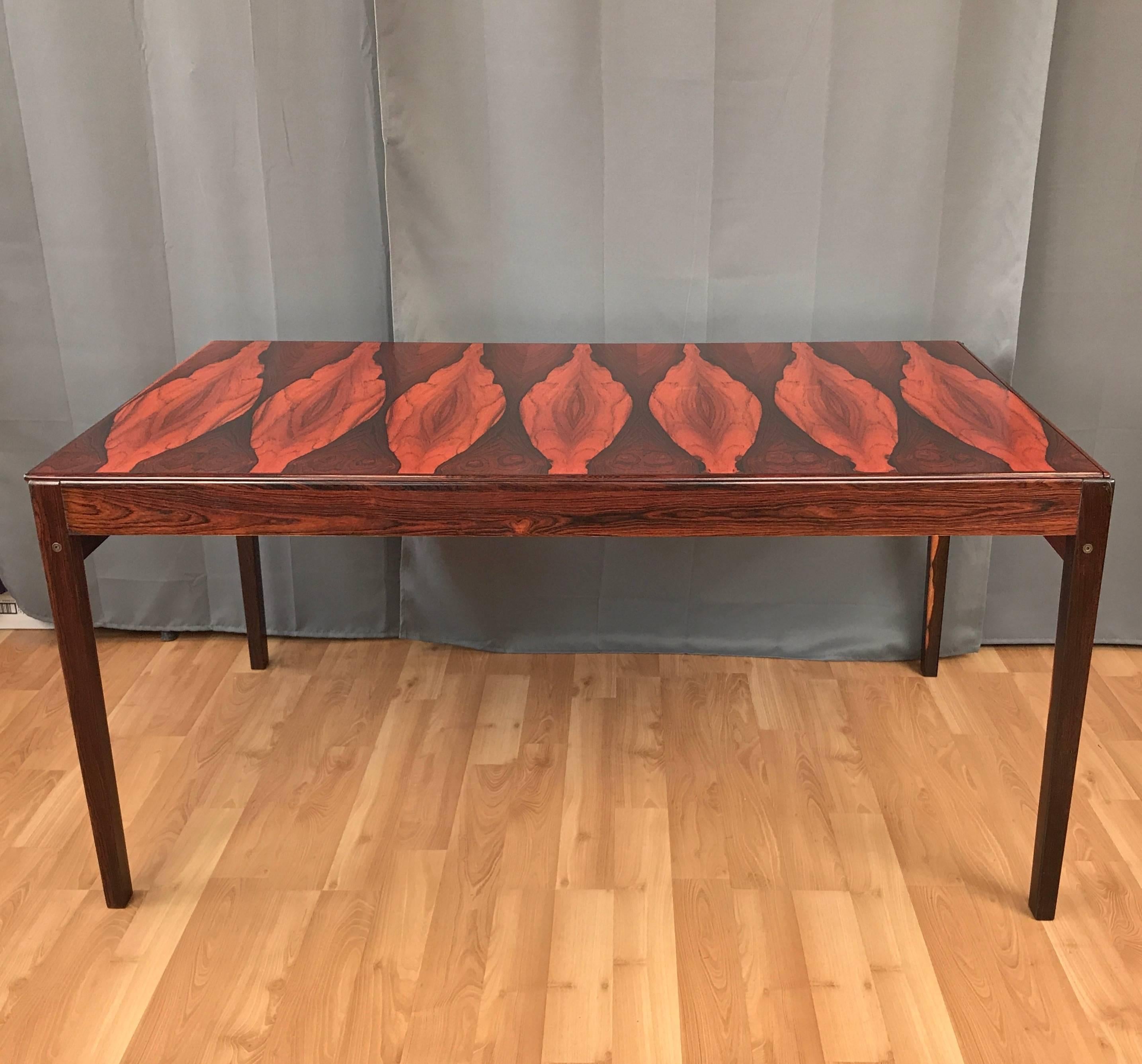 An absolutely dazzling early 1970s Danish expandable rosewood dining table with hidden cartridge leaf. Crotch cut bookmatched veneer top and solid frame display unreal grain, made all the more so by a burnt orange applied tint. Brazilian rosewood
