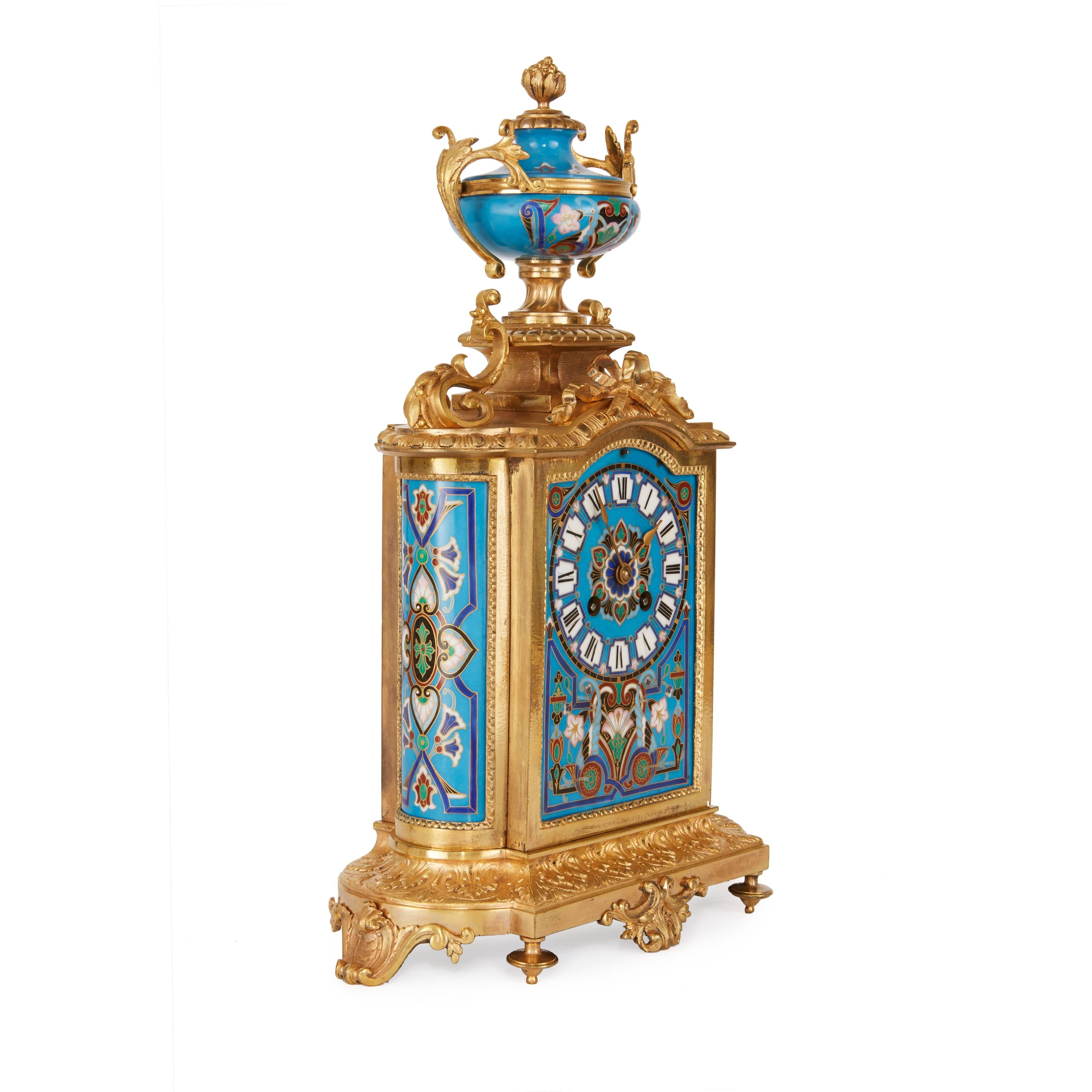 Comprising a central clock and a pair of flanking vases, the clock with central dial set in a panel of porcelain imitating champlevé enamel with geometric design on turquoise ground with an urn surmount, all set on a foliate ormolu breakfront