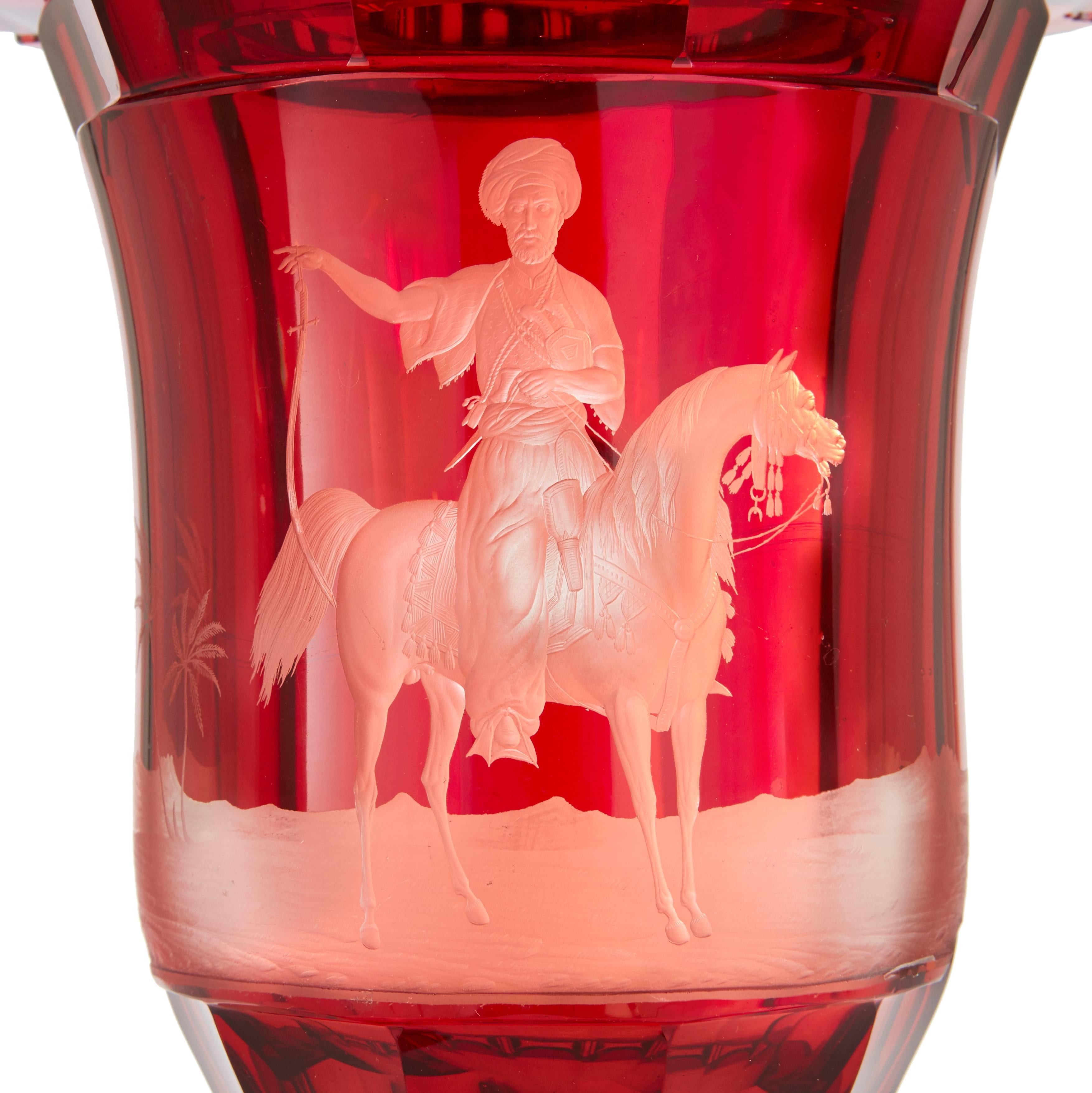 The vase, by Franz Hansel (Czech, 1802-1883), has a ruby glass body that is campana shaped and faceted all around. It is finely engraved to one side with a depiction of a Turkish warrior on his horse. The cover, finial rim and spreading foot are all