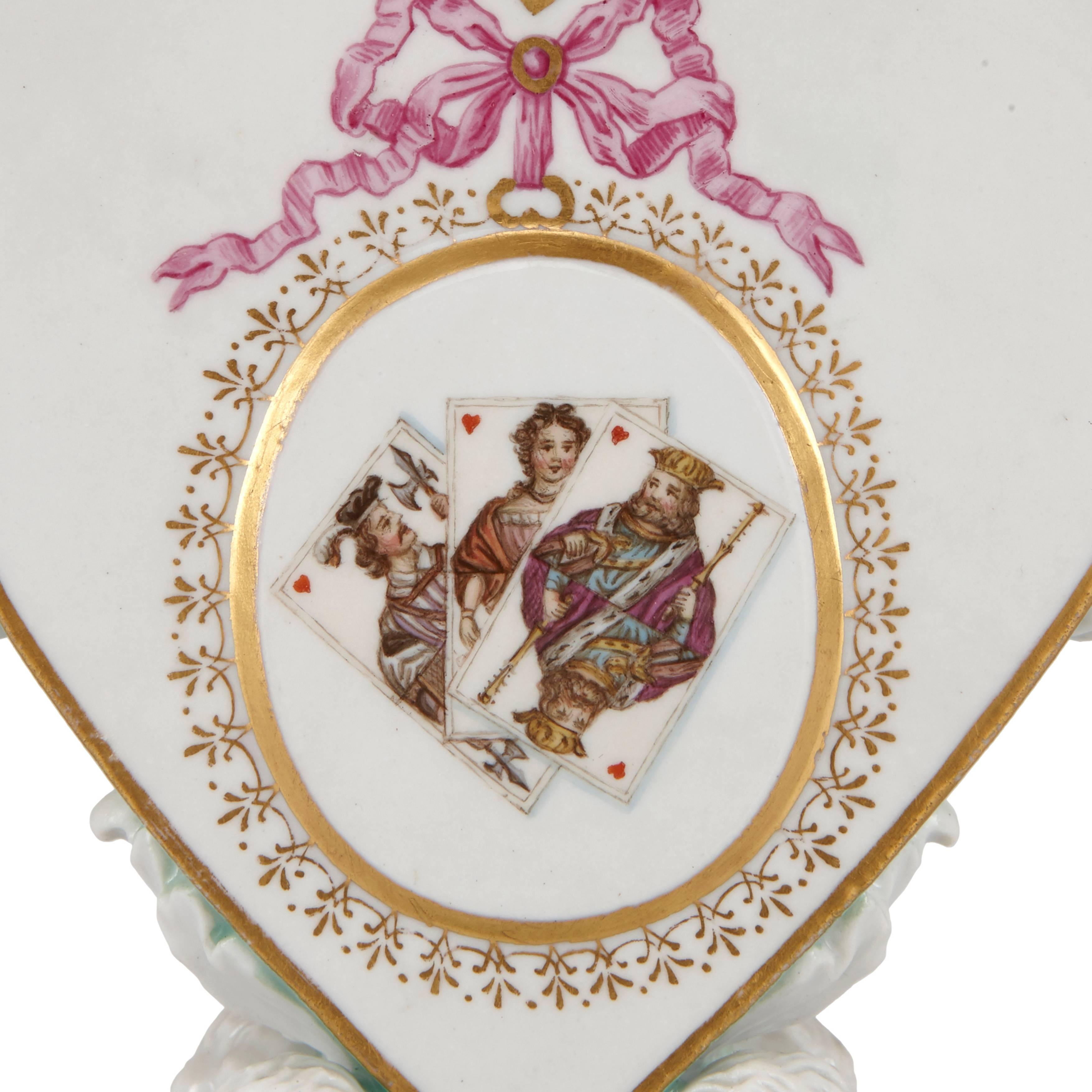 Each candleholder finely detailed to the centre with an image of playing cards to one side and crest on the reverses, each image contained within a gilt circle, further enhanced with gilt pattern work and a pink ribbon, all set within heart shaped