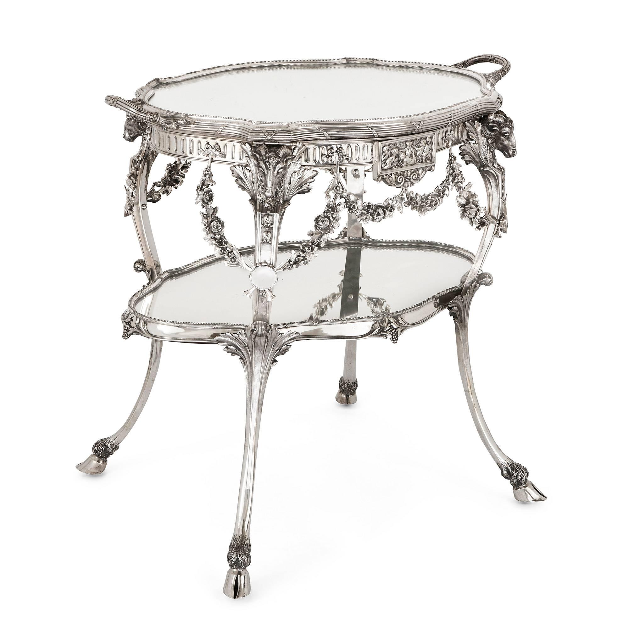 Made of solid silver, raised on four legs topped with ram's head and centred with a carved relief plaque depicting cherubs, the oval shaped tiers surfaced with mirrored glass, the top tier in detachable tray form with beaded rim and twin handles,