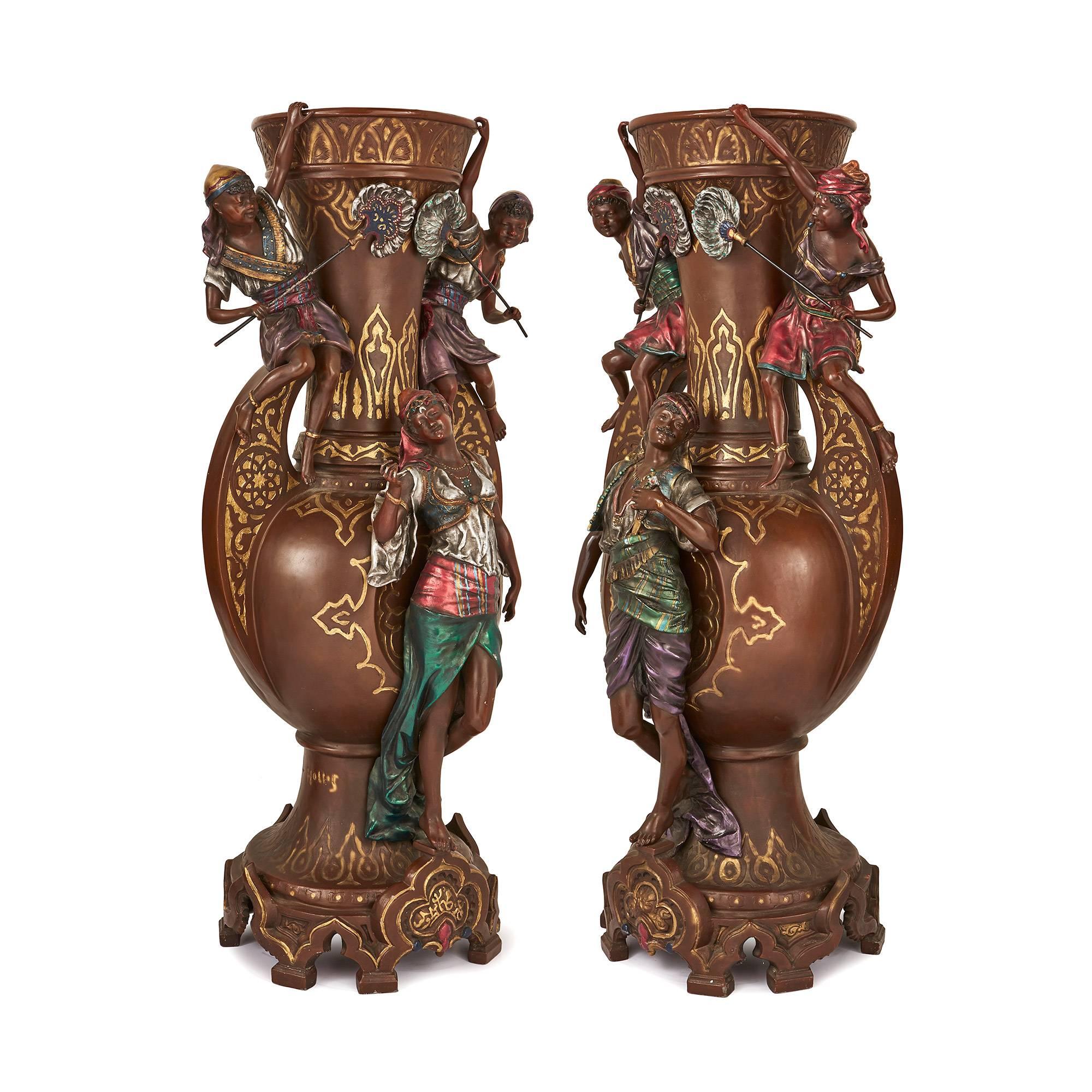 Each of a baluster form in a brown patina and cold painted in polychrome, with an oriental figure in exotic dress, fanned by two servants perched upon the handles on either side with one hand hanging on to the rim, decorated with Islamic-inspired