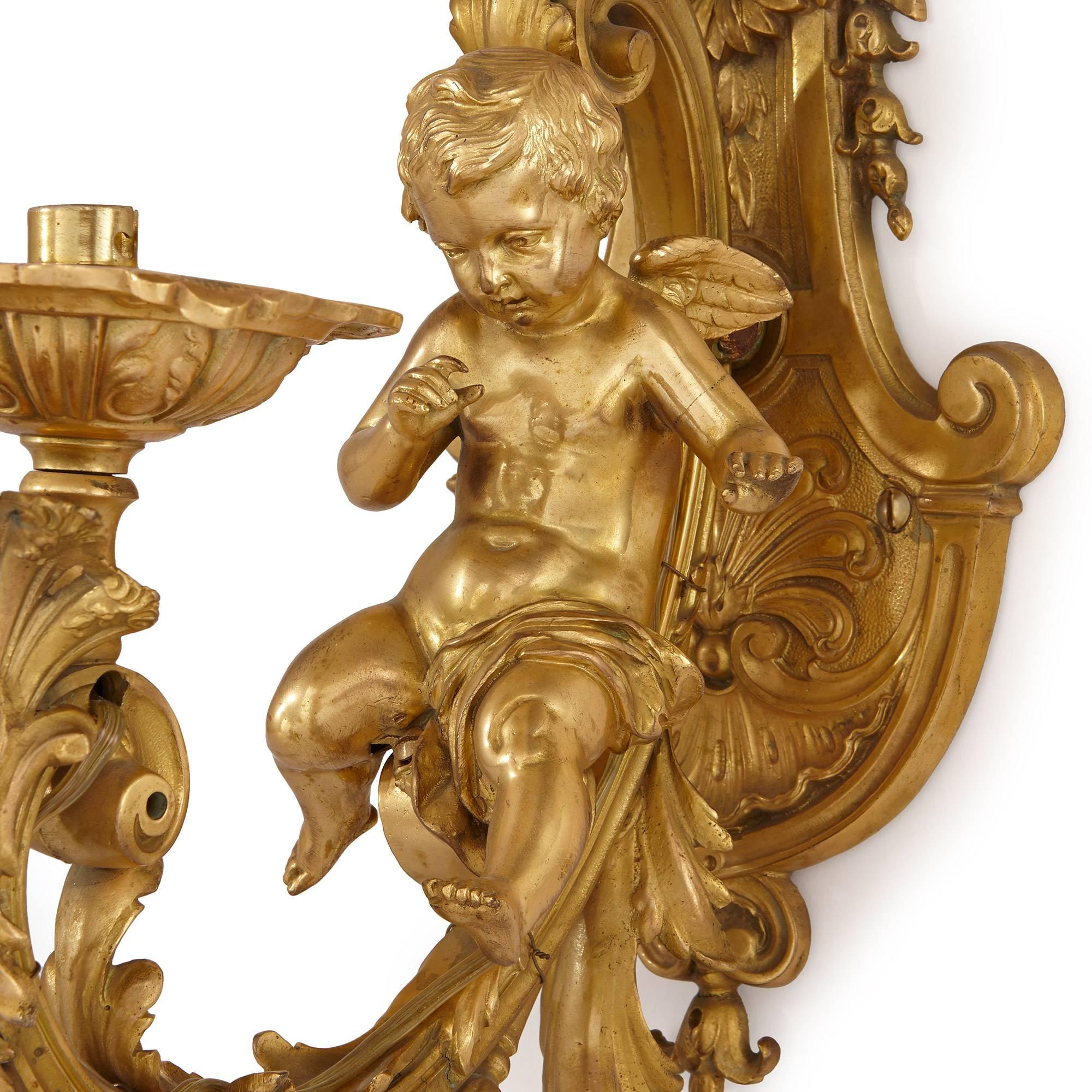 Each with a curved back plate topped with a vase of flowers, a seated putto in front, upon a C-scroll arm ending in a faceted rounded drip plate, mounted in ormolu and heavily adorned with acanthus foliage, floral and scallop edged motifs, signed