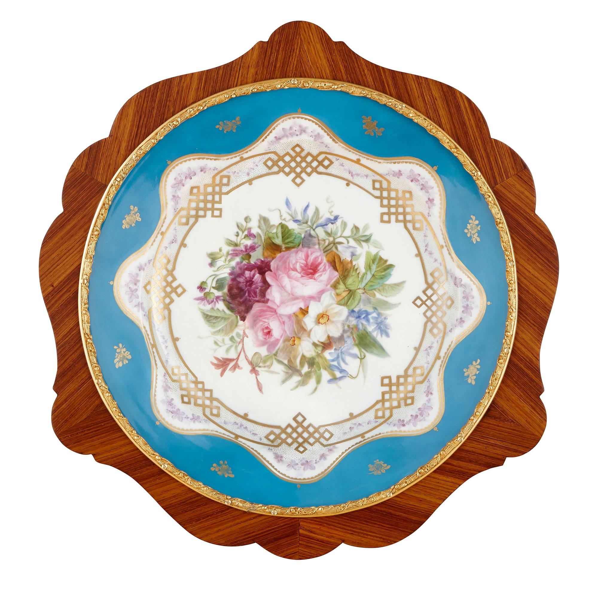 Set with a large central circular turquoise ground painted porcelain plaque depicting flowers, the wooden frame set with a beaded ormolu border and further scrolling ormolu decorations, leading to three scrolling legs joined in the centre.

This