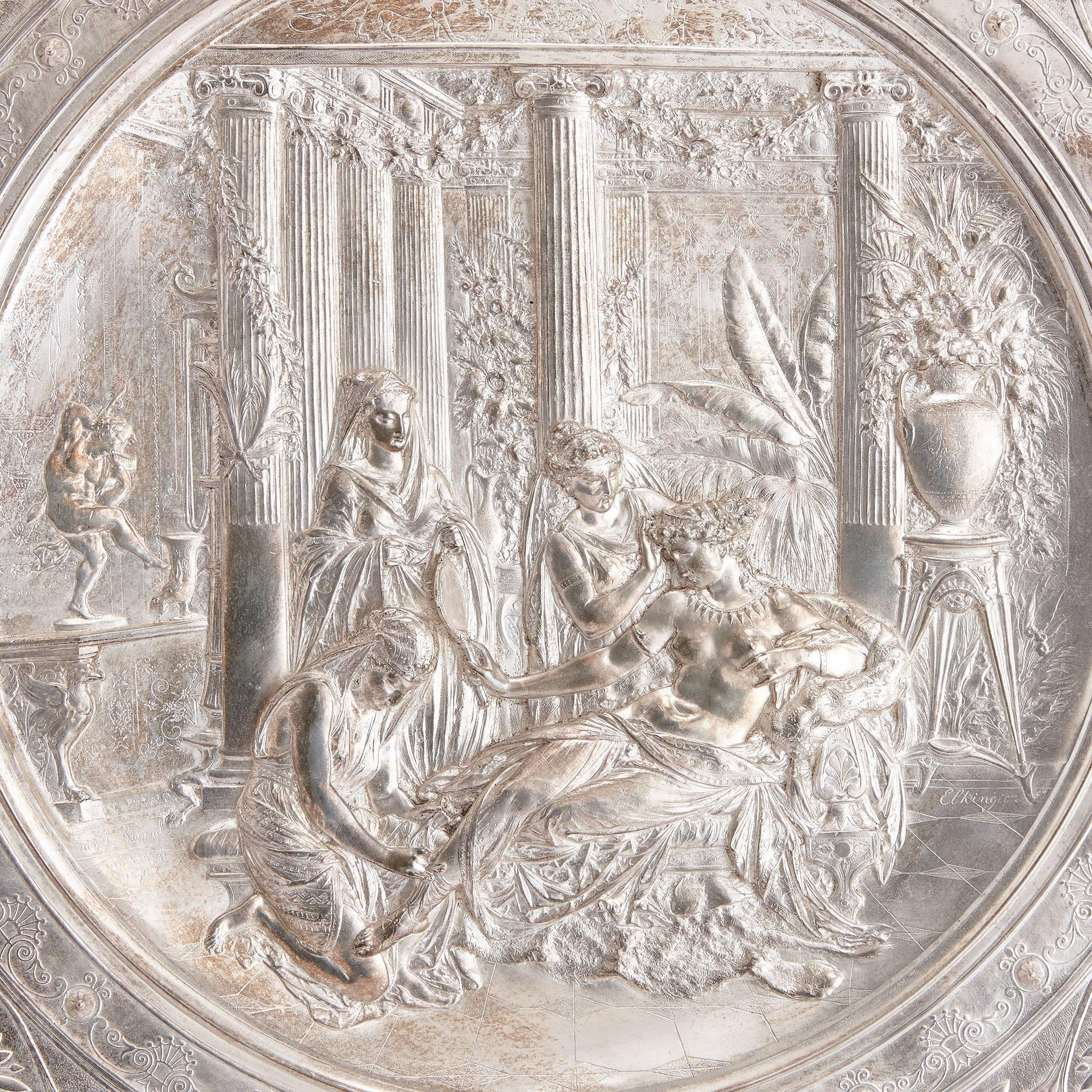 The silver charger mounted on a velvet board, of circular form in the Neo-Grec manner, depicting a classical figural scene of a semi-clad maiden surrounded by three female attendants with an exotic colonnaded interior, within a florally decorated