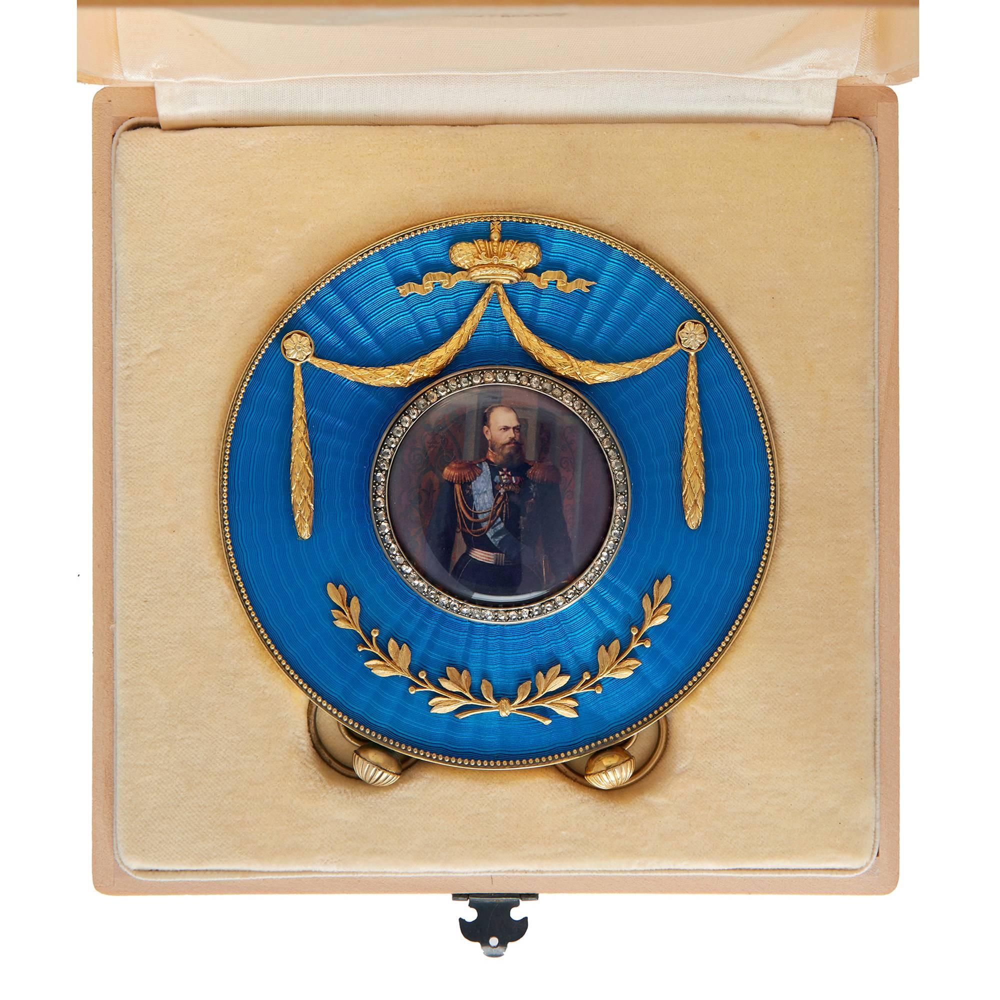 The central circular photograph surrounded by a band of diamonds, set on a circular blue guilloché enamel panel surmounted by a crown and with floral decoration within a silver gilt border and easel backing, with spurious hallmarks and fitted wooden