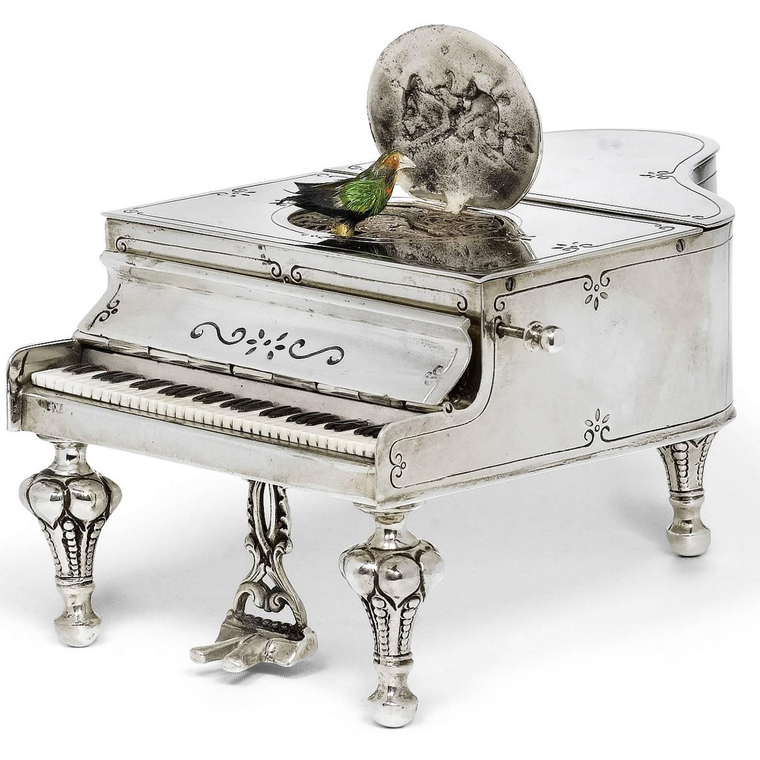 This charming antique silver musical box features a singing bird, which appears from an opening oval. The musical box is exquisitely crafted in the shape of a miniature grand piano, featuring a hinged medallion with emblems, a closing keyboard cover