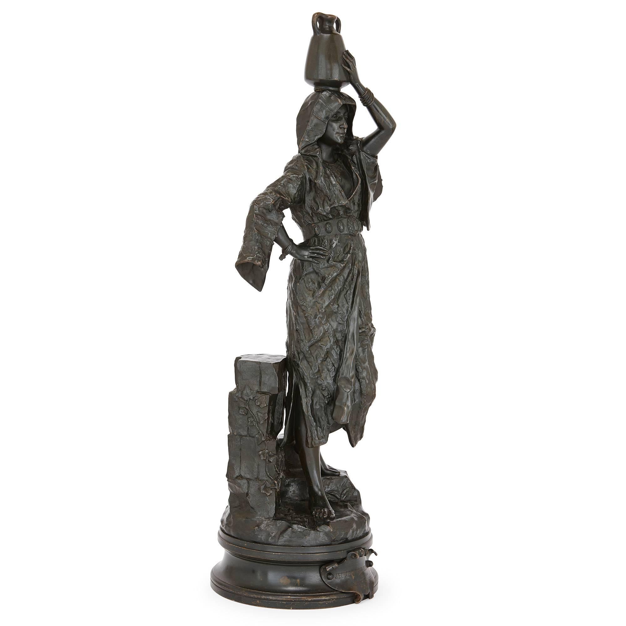 This charming sculpture depicts a young Arabic girl, in typical dress, carrying a jug of water on her head, standing on a natural base and all set on a circular plinth with title plaque. It was created by the French artist Gaston Leroux who was