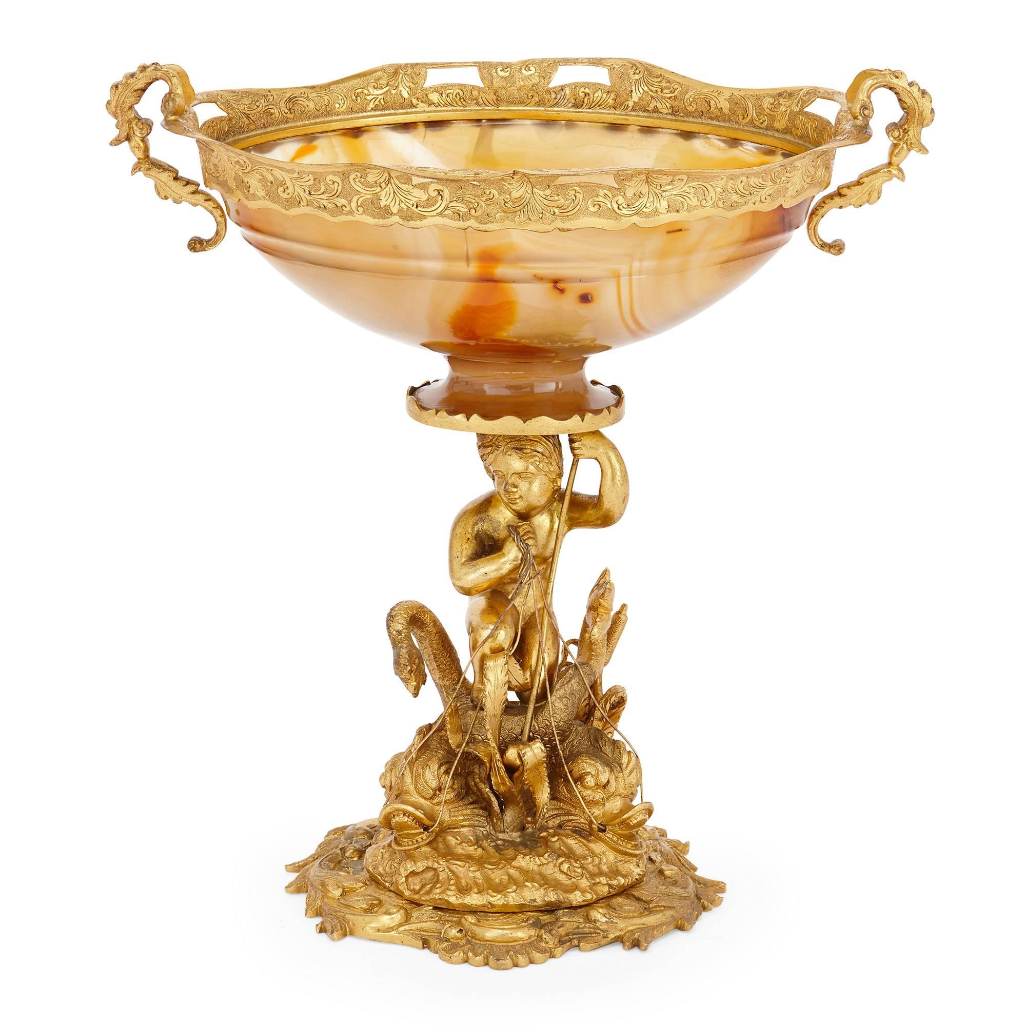 This fine ormolu centrepiece is modeled as a young boy standing on sea creatures from classical mythology, all set on a naturalistic base. A bowl made from agate, the orange and yellow coloured crystalline rock material, sits on top of the base and