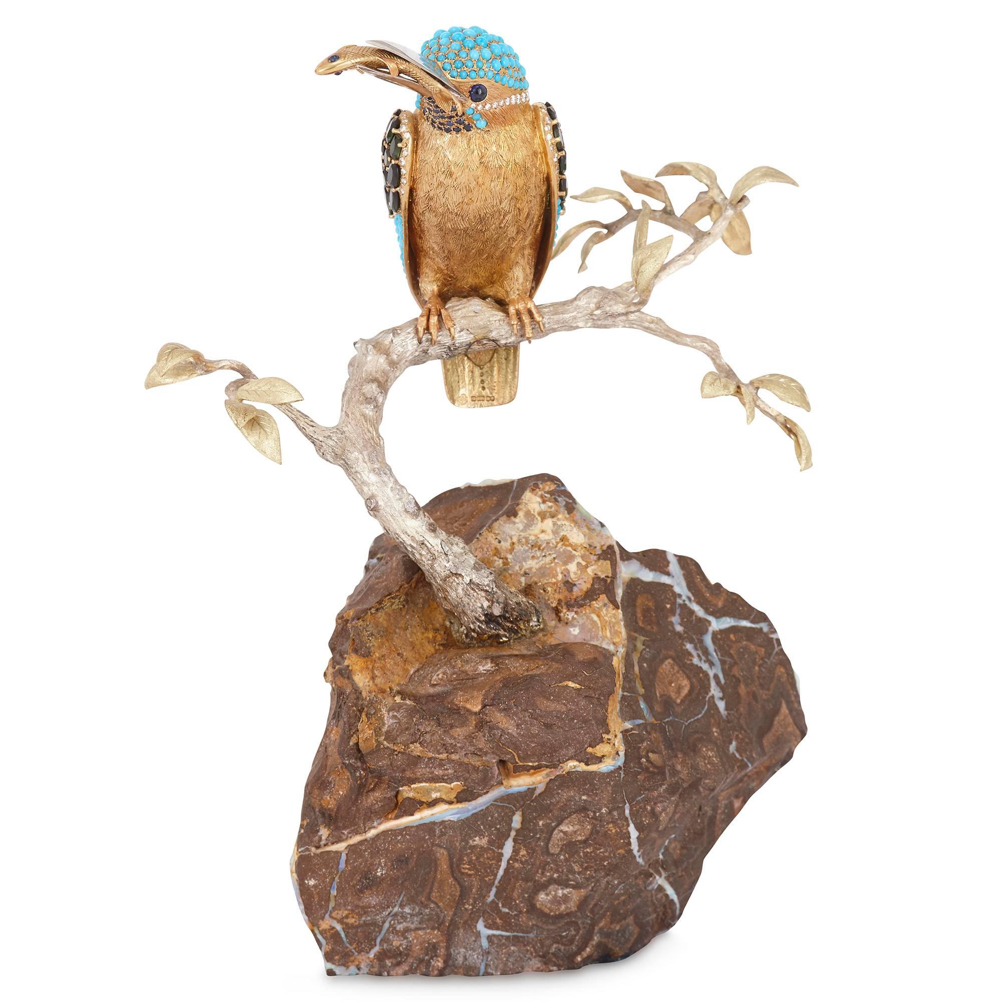 Beautiful gold model of a kingfisher set with precious gemstones including sapphires, diamonds, turquoises and peridots, all set on an opal boulder base. The gold bird is perching on a tree branch, holding a gold fish in its beak. The underside of