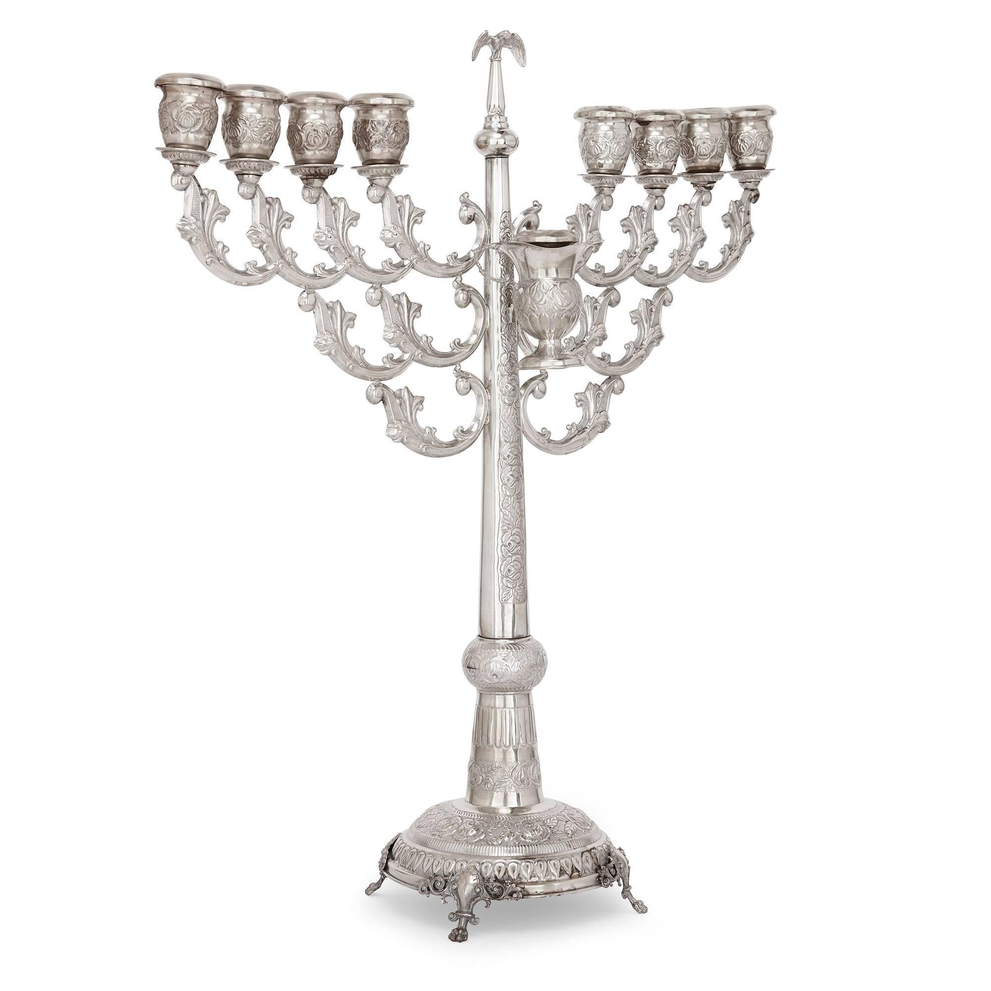 This large antique silver menorah features a central column with fine detailing using the repousse technique, surmounted by an eagle finial which is able to convert to the shammash, the ninth candle holder. A removable silver oil jug sits below,