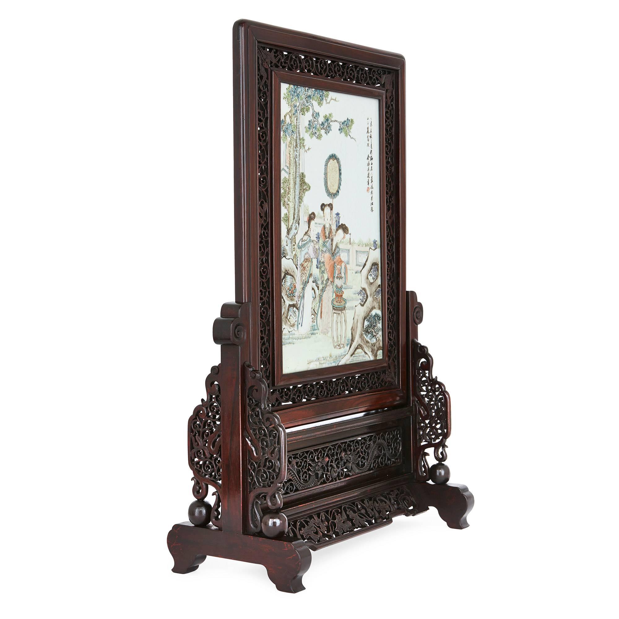 This exquisite Chinese screen is crafted from Hongmu, a dark rosewood from South East Asia. The ornately carved frame depicts dragons and scrolling foliate shapes. The central porcelain plaque depicts Chinese ladies in a detailed landscape with a