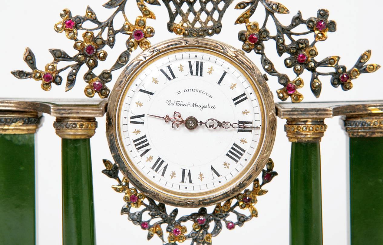 This delicate table clock features a central circular white dial with black Roman numerals and gilt detailing. The dial is signed 'E. DREYFOUS, To Their Majesties' and is mounted all around with silver worked flowers and a crown motif, all inlaid