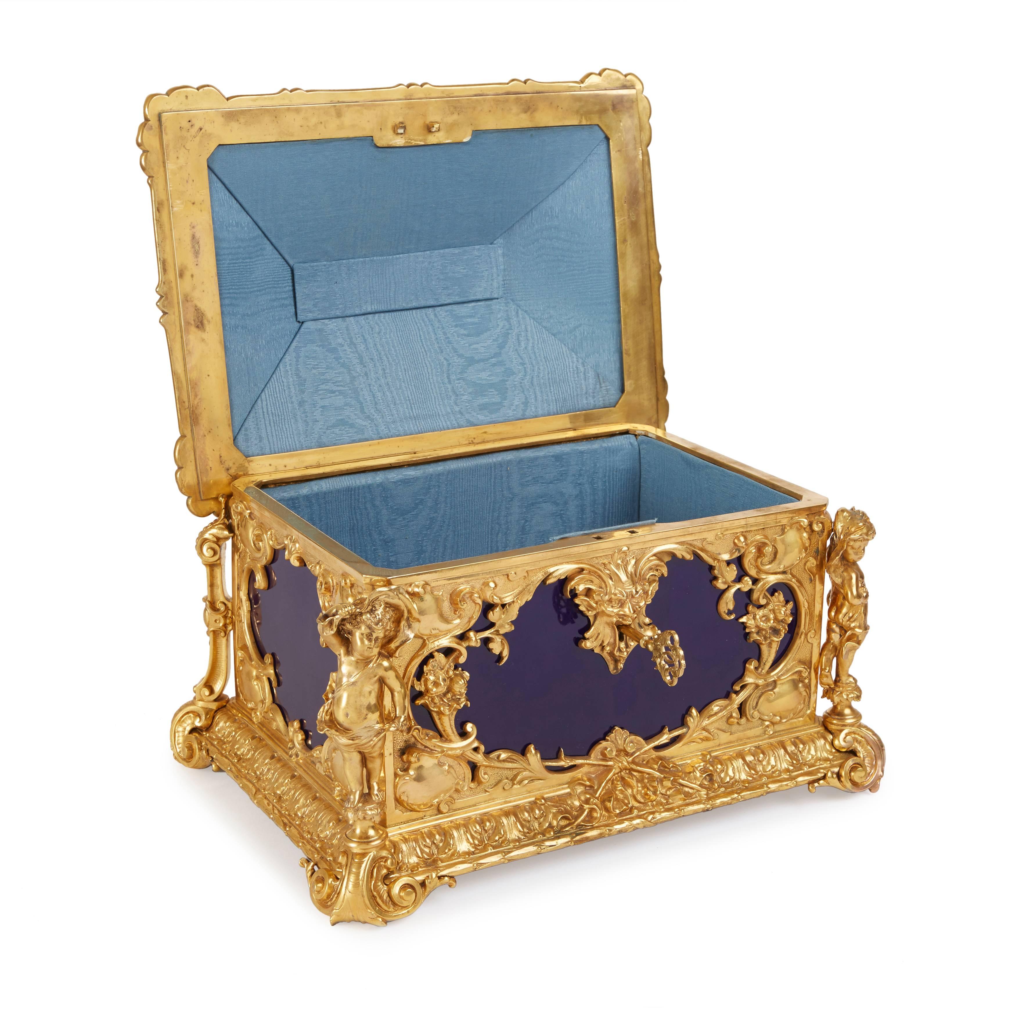Of rectangular form, the cobalt-blue ground porcelain plaques mounted with rich ormolu corner mounts with scrolls, floral bouquets and a grotesque mask escutcheon, the hinged cover topped with an ormolu cherub handle, on scroll feet, the porcelain
