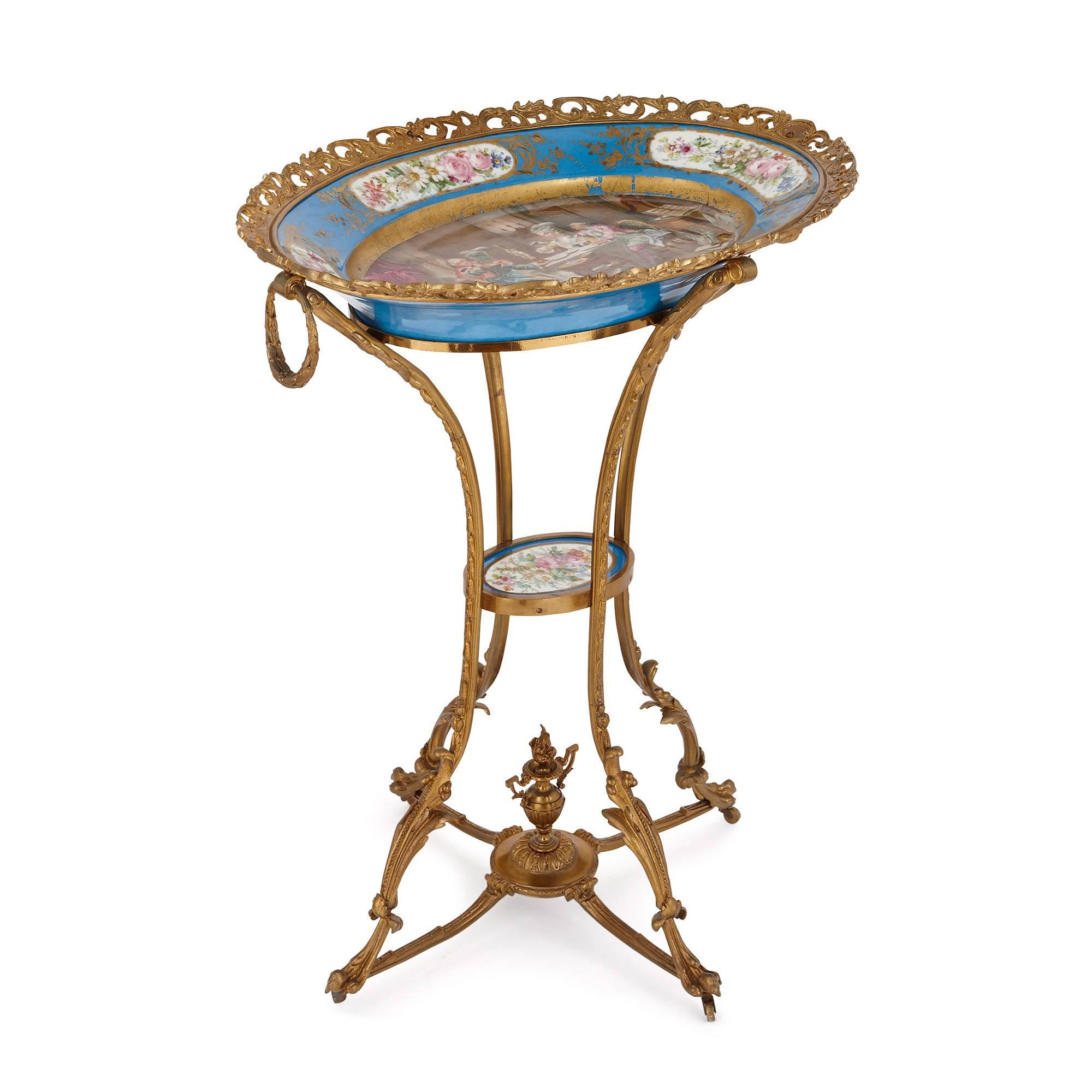 The oval shaped porcelain charger depicting 18th Century aristocratic men and women sat at a table with a floral pattern rim and a pierced ormolu frame, set on four ormolu legs joined in the middle with a porcelain plaque.

This stunning Louis XVI