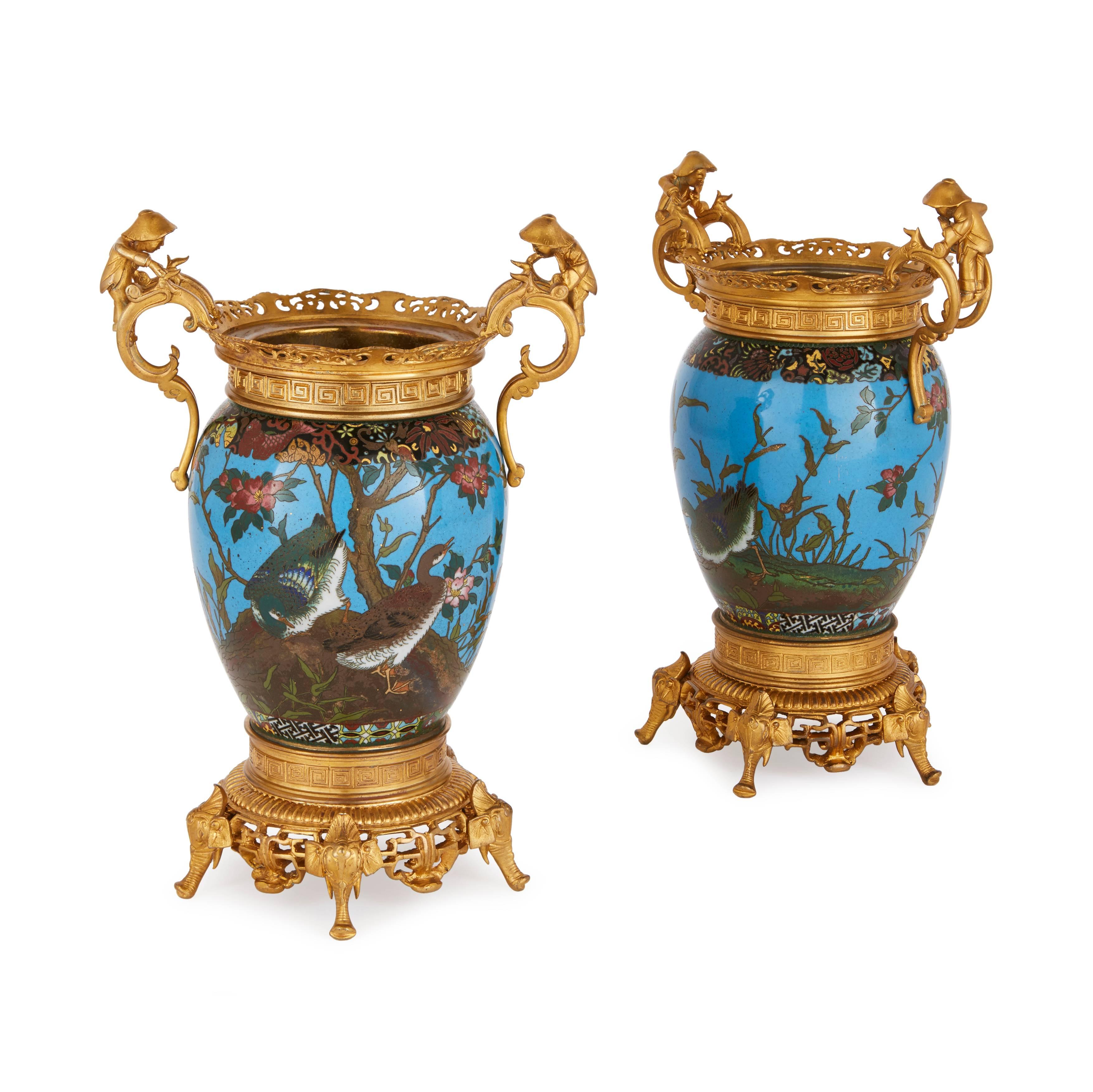 The pair of ovoid form with a pierced ormolu rim and scrolling handles modeled with Japanese figures, decorated throughout with fowl, flowers and foliage, raised on elephant feet. The Japanese enamel vases dating from the Meiji period, with French