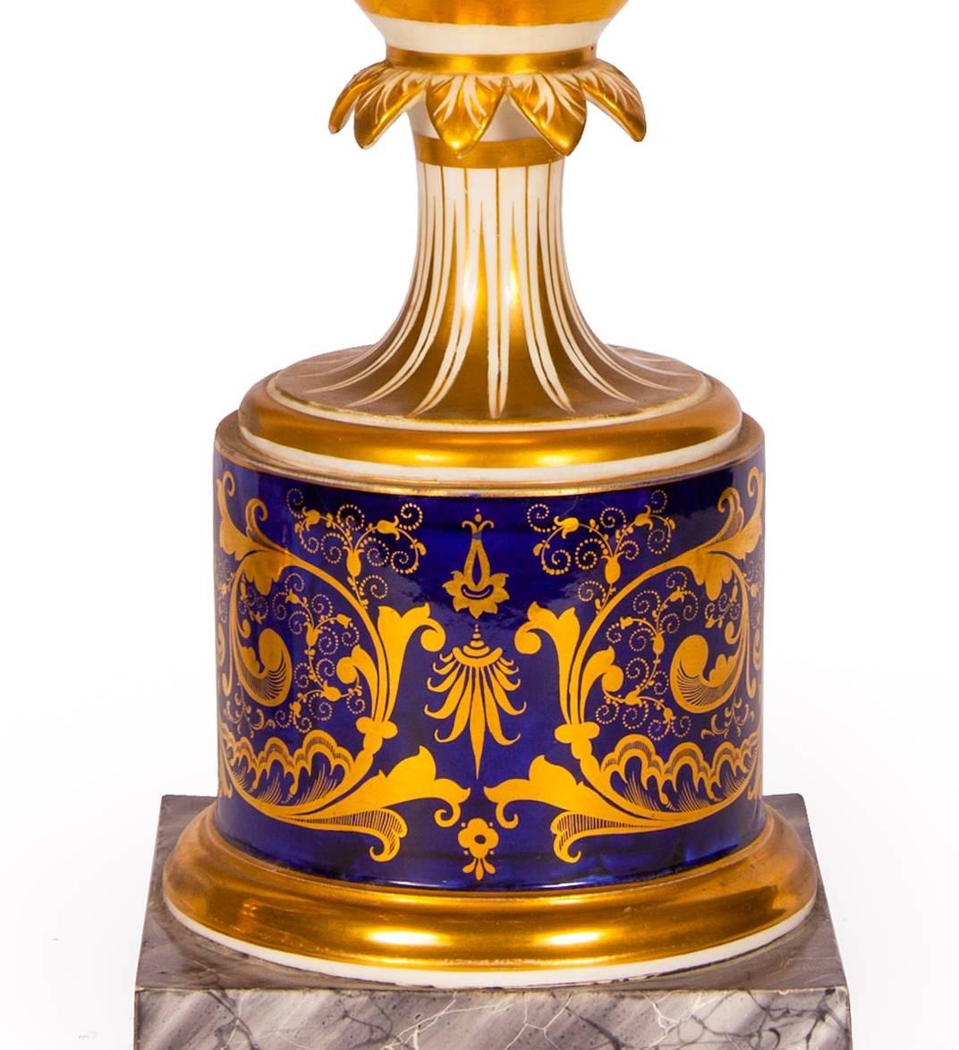 The pair of urns are each mounted on square plinths of grey veined marble. The lower sections of each of the urns are cylindrical and are decorated with gilt ground arabesque patterns on a blue ground. The bodies of the urns are of ovoid form, and