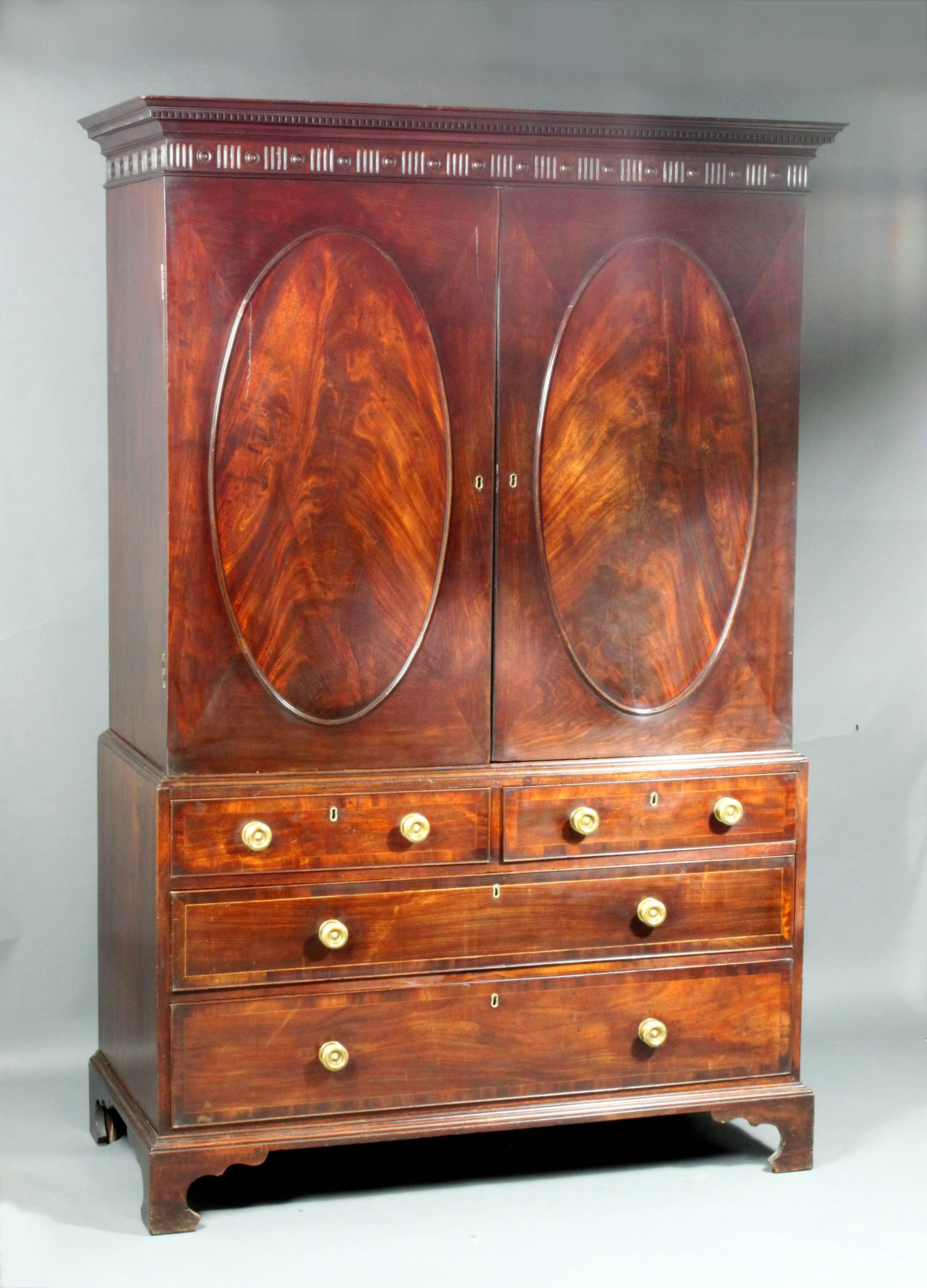 A fine George III Sheraton period oval panelled mahogany linen press of a good color and patina: The cupboard with three cedar lined slides and oak lined drawers below, fine carved dentil cornice. Replaced brasses.

Cornice measurements: 52 1/4