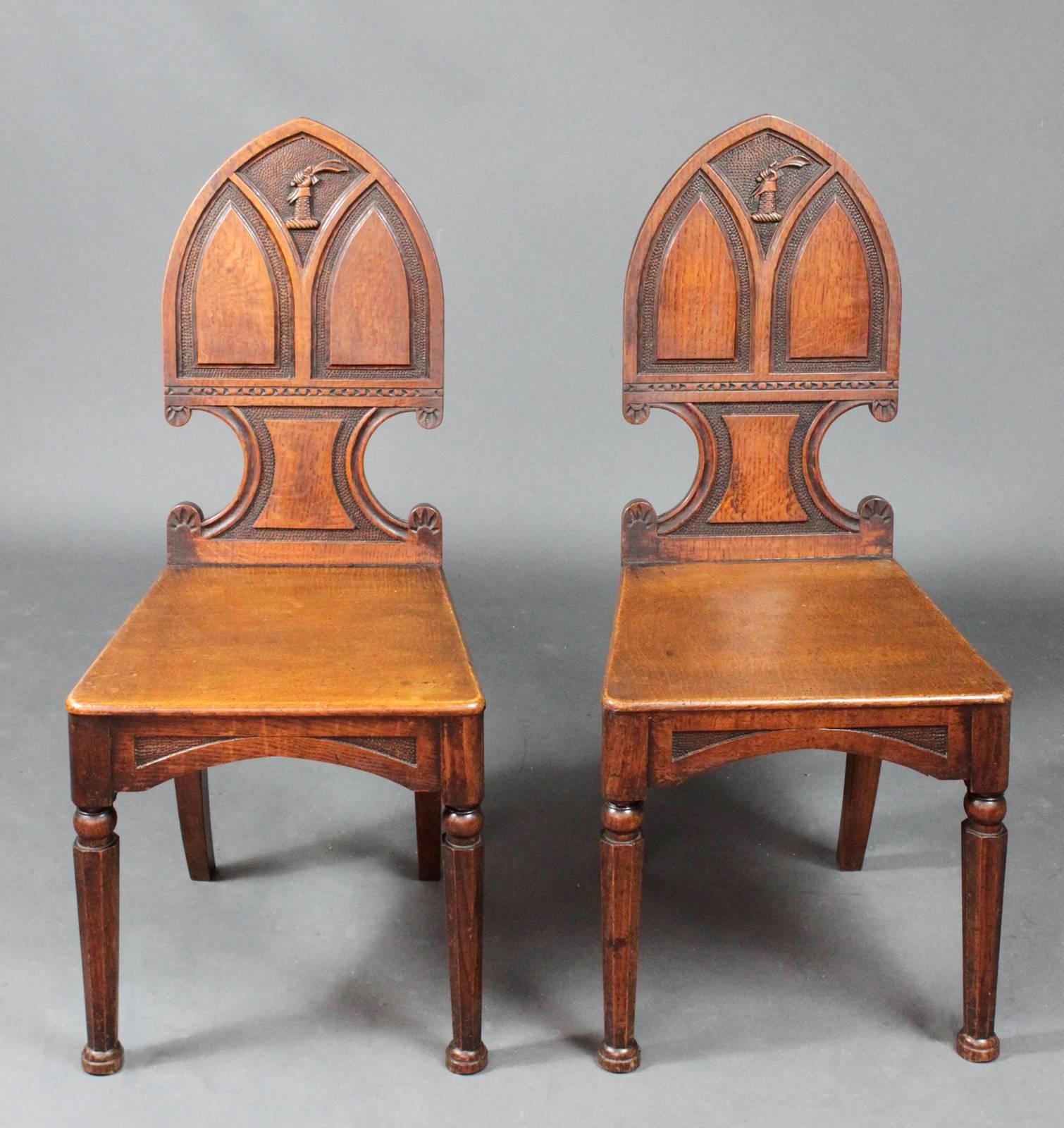 A good pair of oak Gothic hall chairs with a well carved family crest of a gauntletted arm holding a sabre aloft. Original condition.