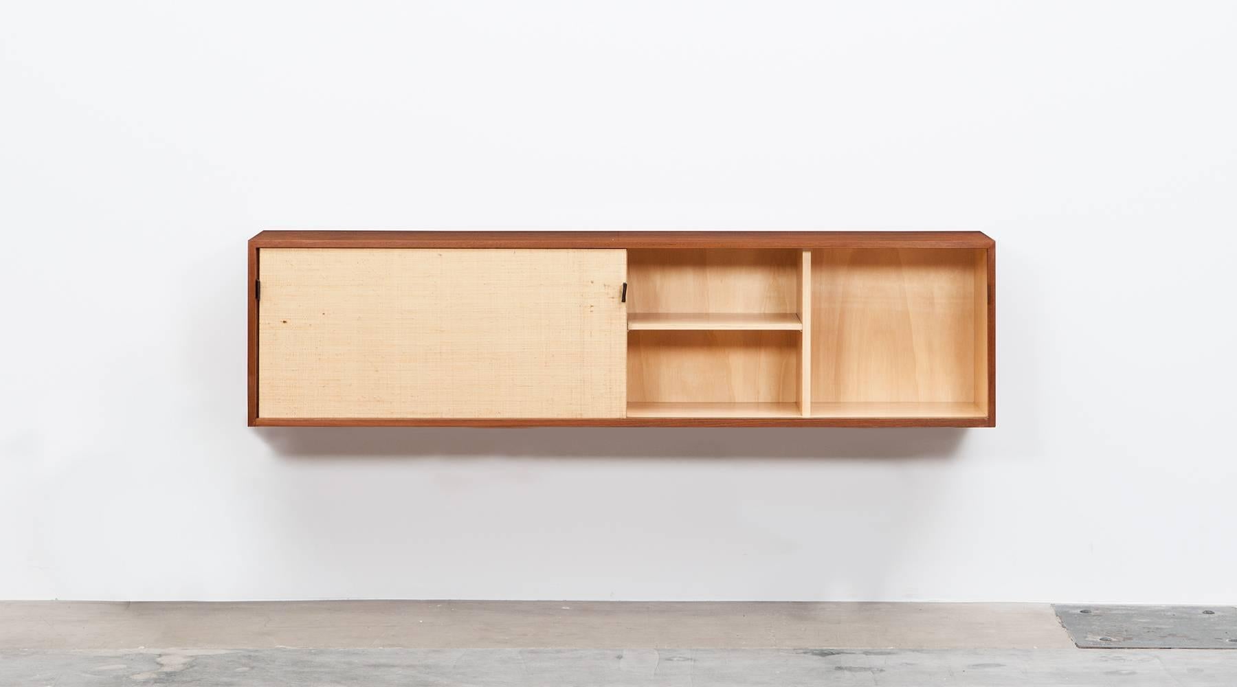 A very nice rarely example of a wall-mounted sideboard in teak designed by Florence Knoll. This model is with two sliding doors covered with seagras paper and leather pulls. The inlay is equipped with shelves for a lot of storage space. We have